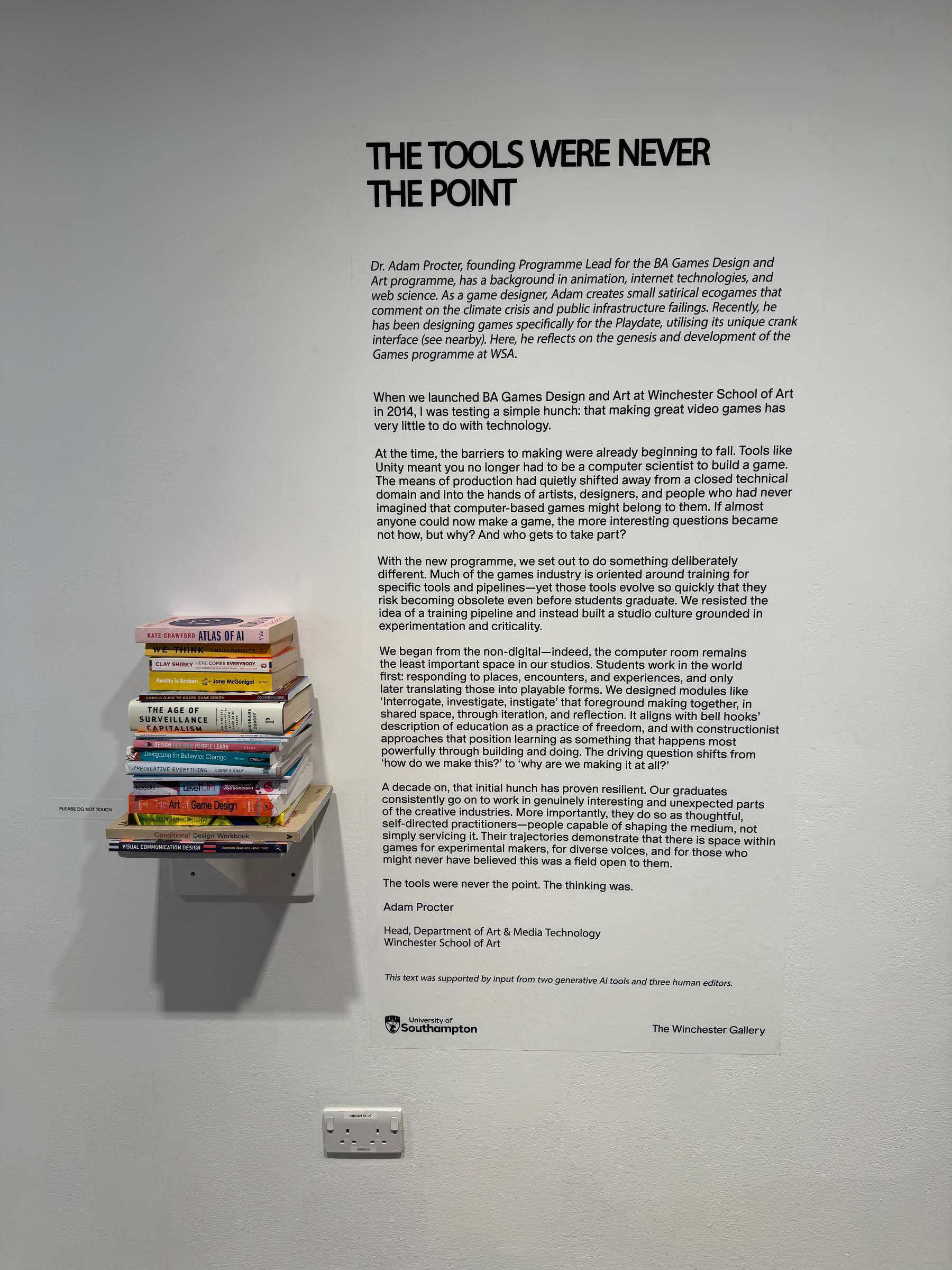 A stack of books is displayed on a small wall shelf next to a large wall text about the history and development of BA Games Design and Art at Winchester School of Art.
