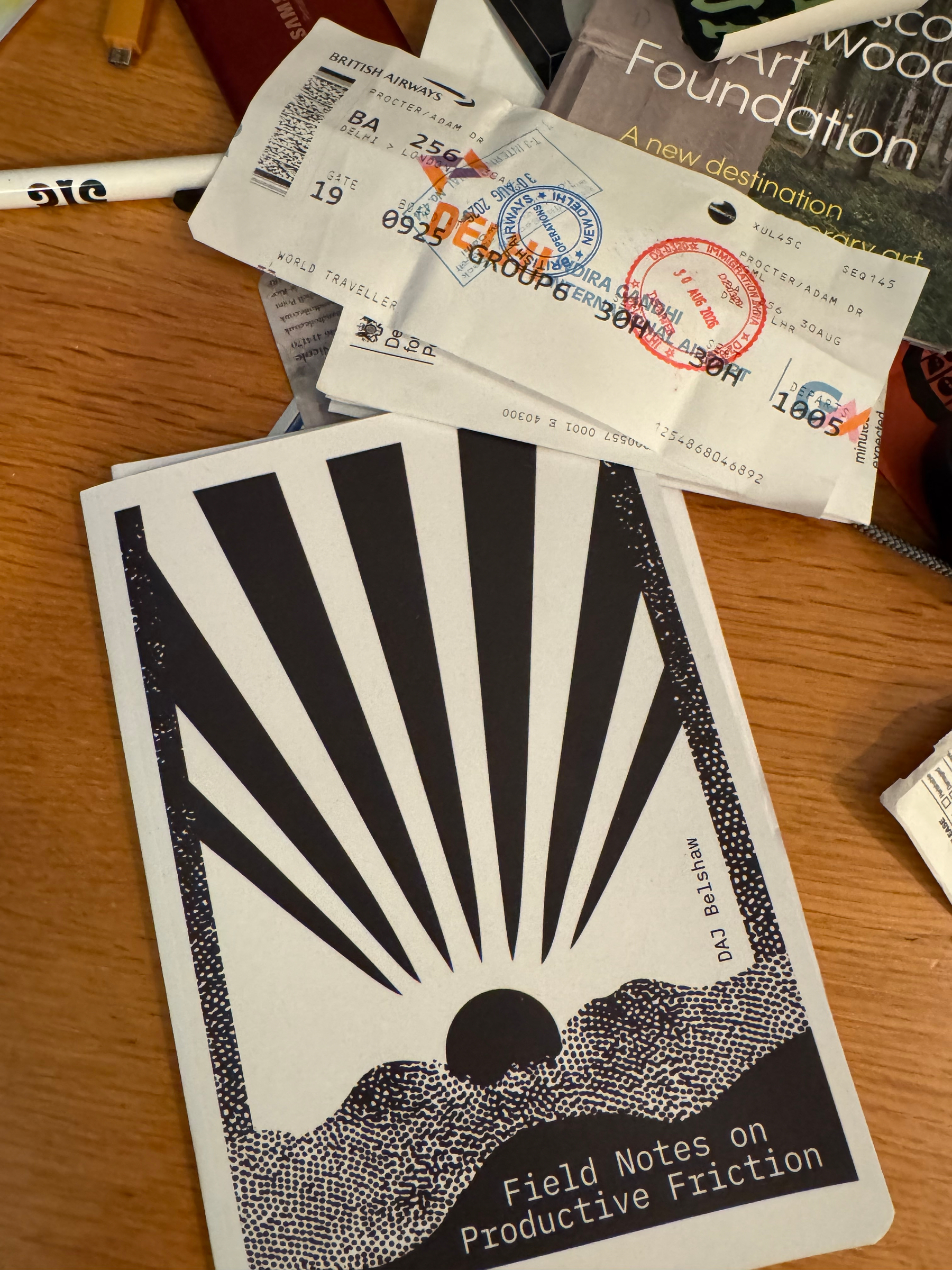 A notebook titled Field Notes on Productive Friction with a stylized sun and rays cover design is placed on a wooden surface alongside some old airline boarding passes and magazines.