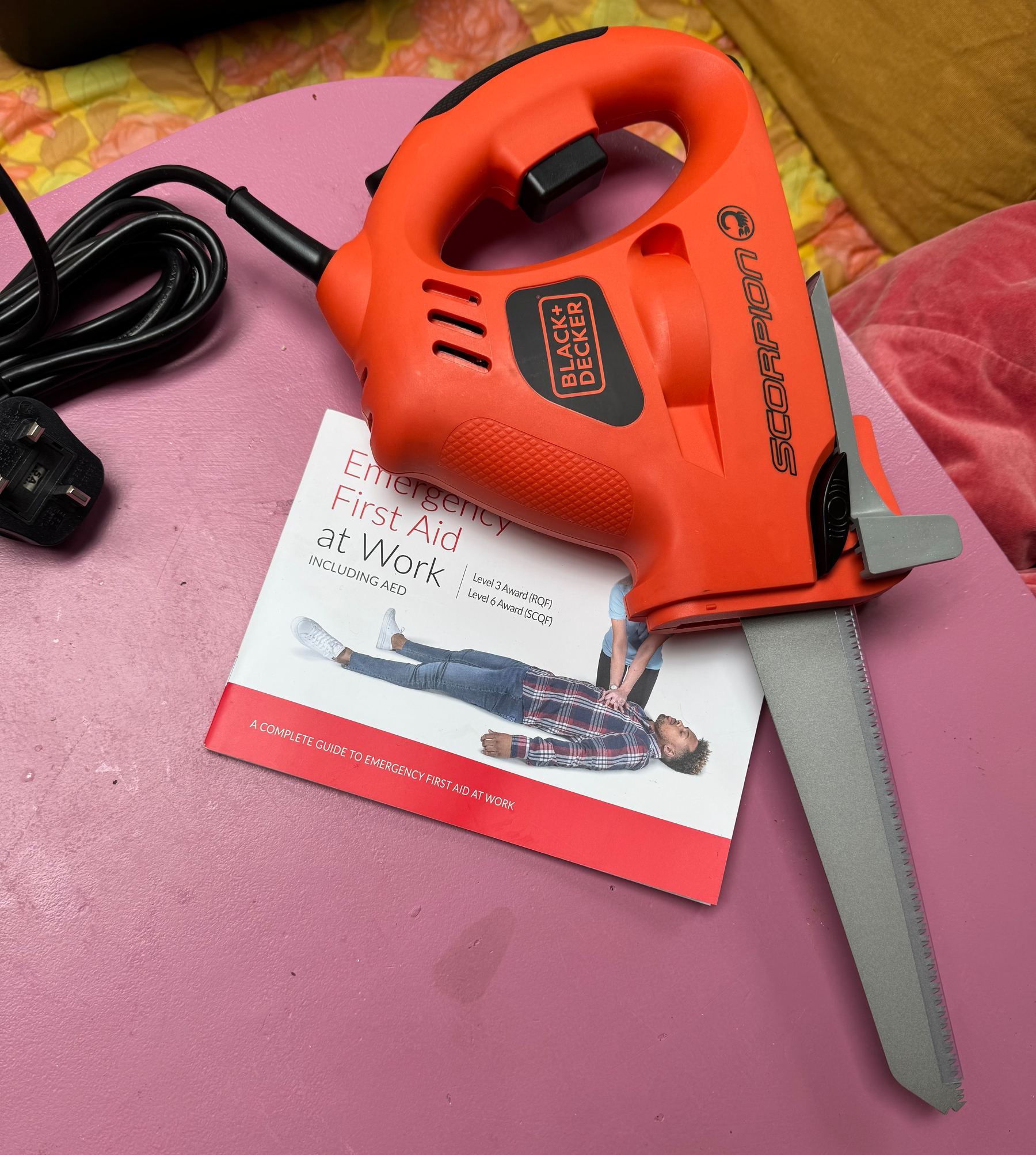 A red Black & Decker Scorpion electric saw is placed on a pink table next to an Emergency First Aid at Work booklet.