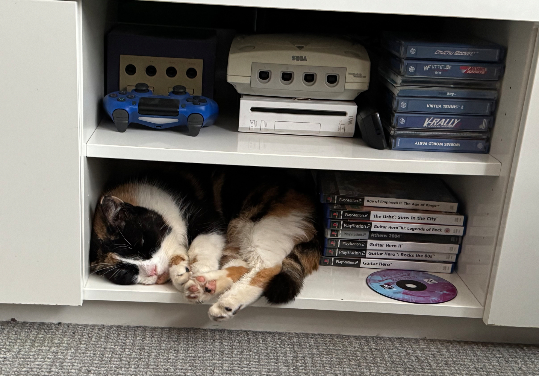 A calico cat is sleeping curled up inside a shelf next to video game consoles, controllers, and game cases.