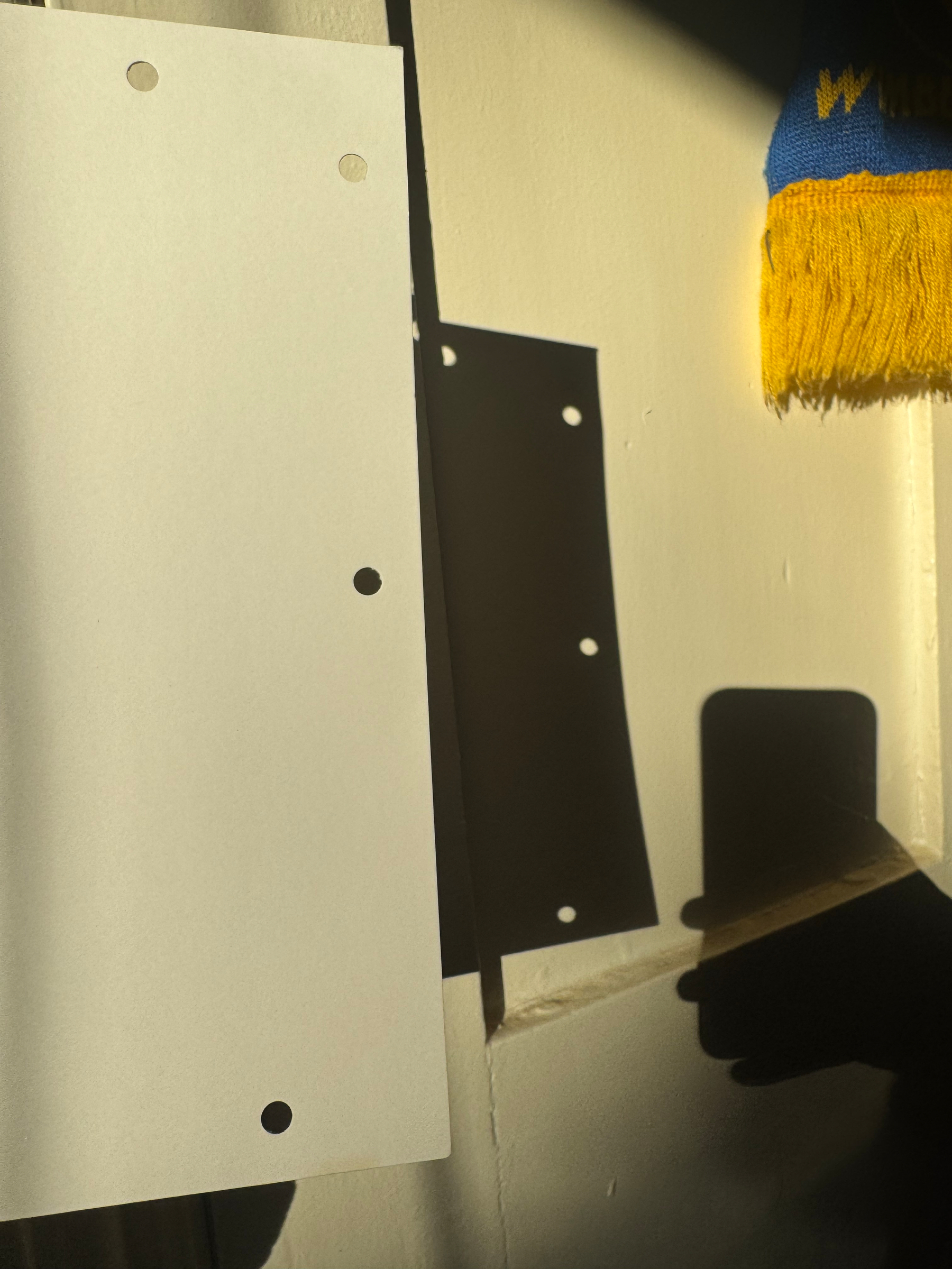 A sheet of paper with holes punched in it is held up by a hand, casting a shadow on the wall along with the hand holding a phone and a colorful textile with yellow tassels from an AFC Wimbledon scarf hanging nearby.