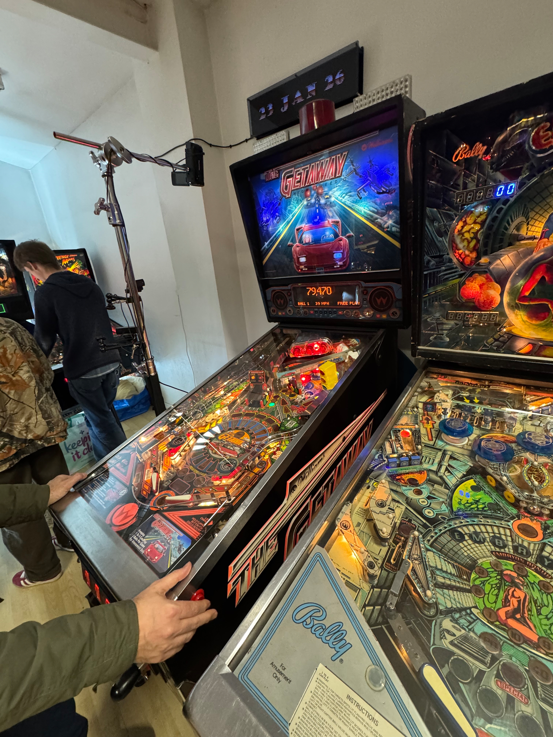 Two people are playing on pinball machines at an arcade, with one machine displaying a GETAWAY theme.