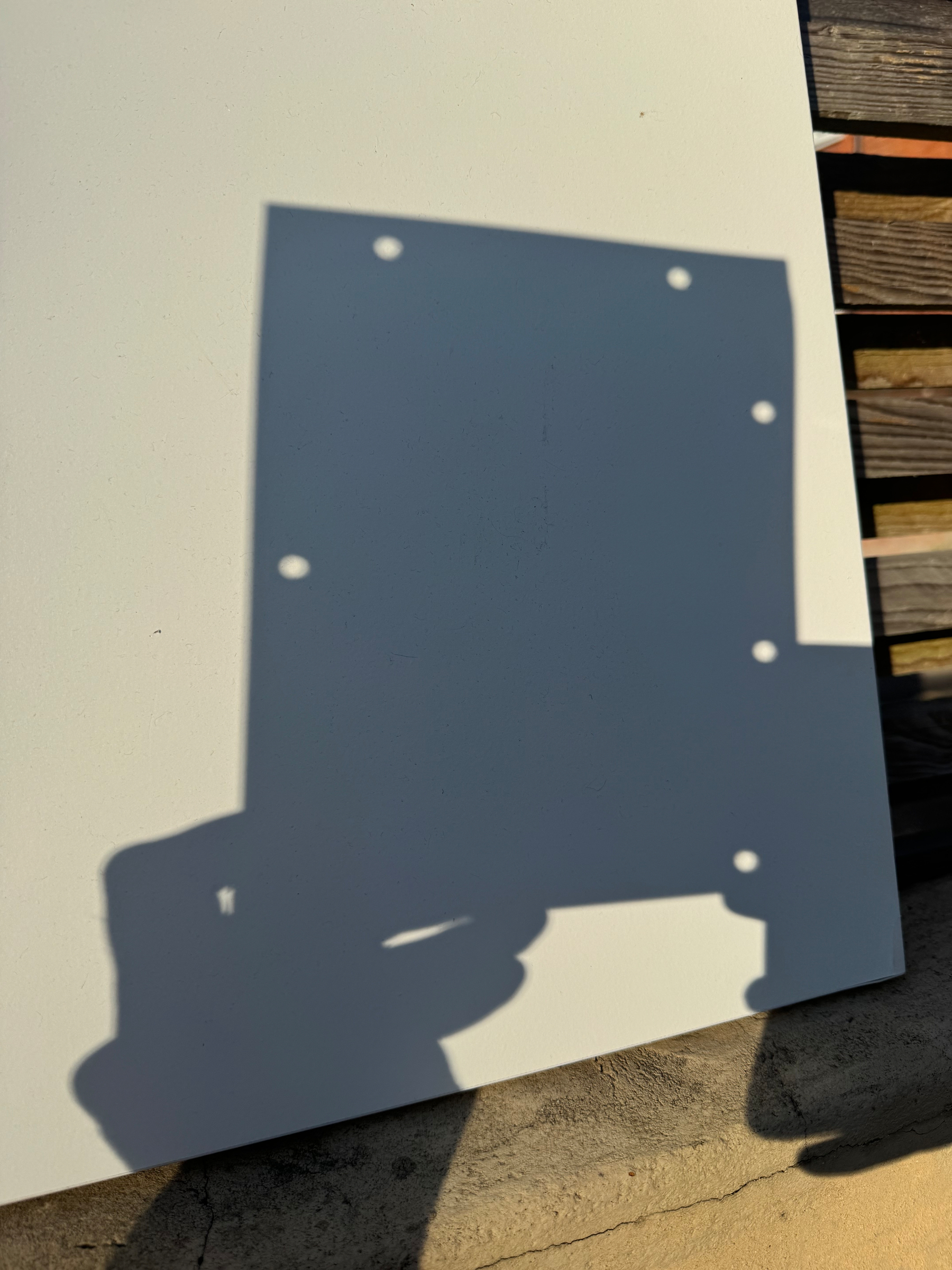 A shadow of a rectangular object with multiple holes is cast on a white surface next to a wooden plank.