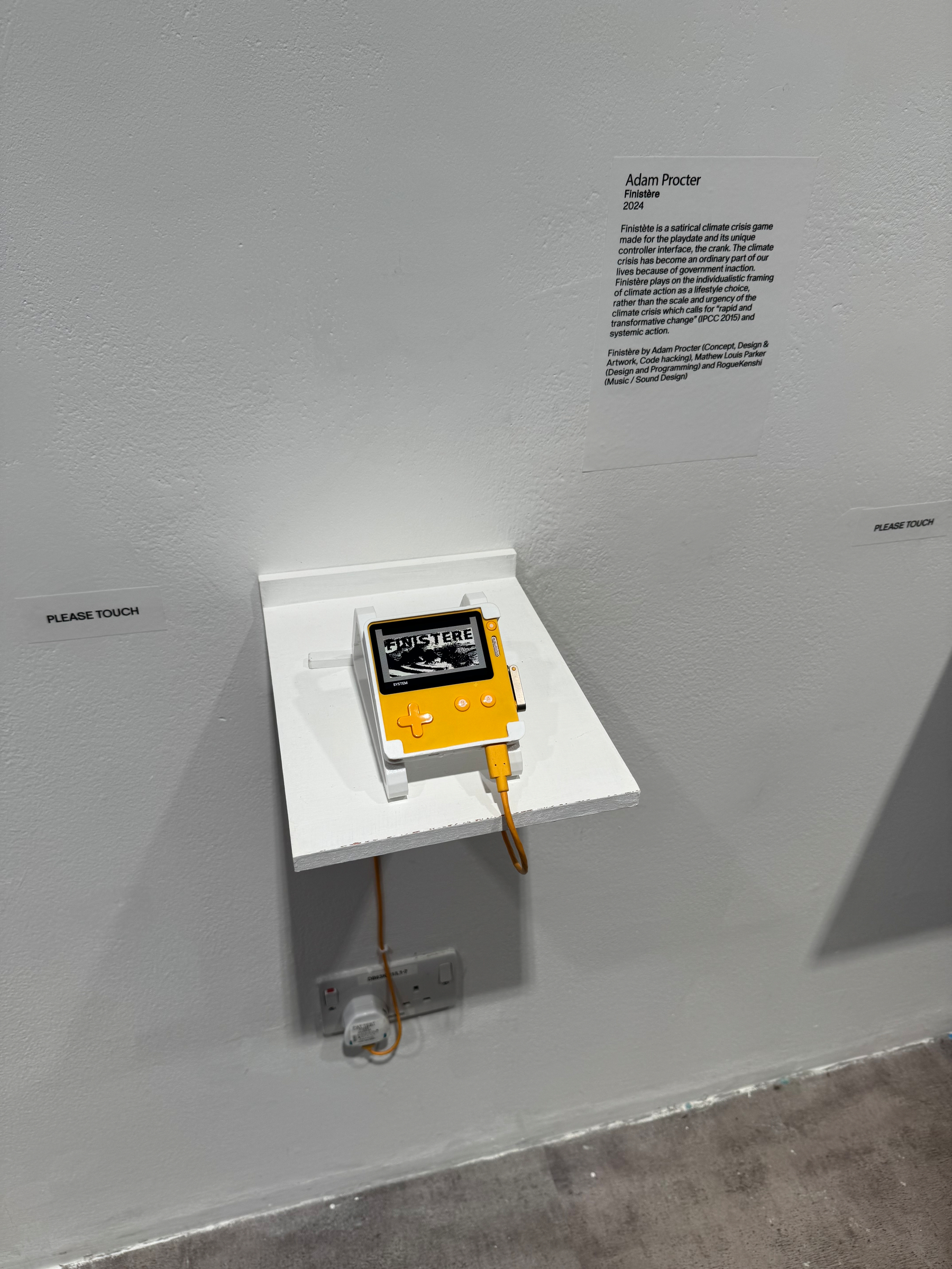 A small gaming device with an orange cable is displayed on a white shelf, accompanied by a description on the wall.