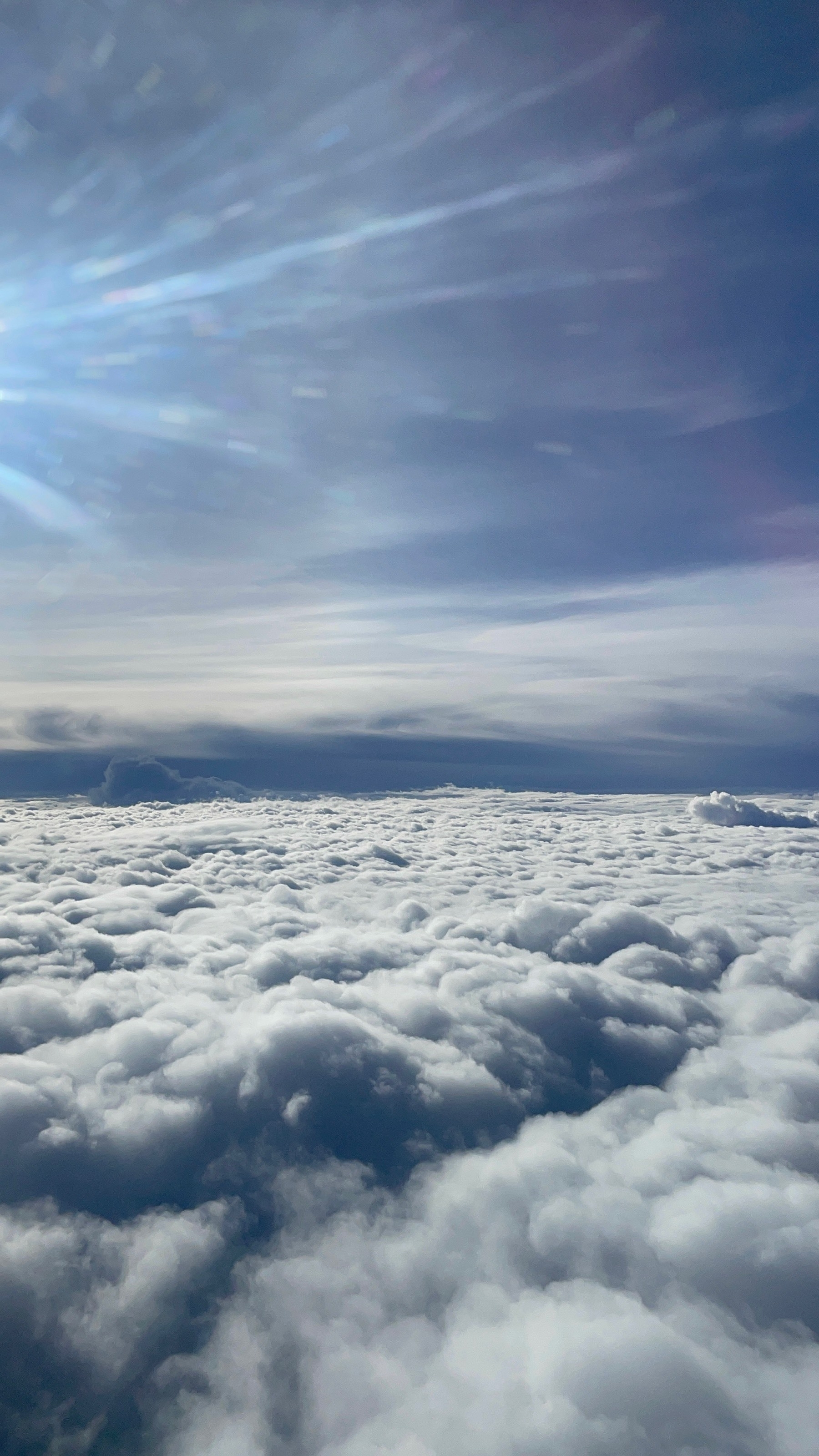 View outside a flight en route to Thailand from the Philippines. The glare of the sun is out of the frame, but its light shines brightly on the fluffy sea of clouds, further defining their shape. The sky above the clouds is a calming type of blue, reinforcing how serene the heavens look.