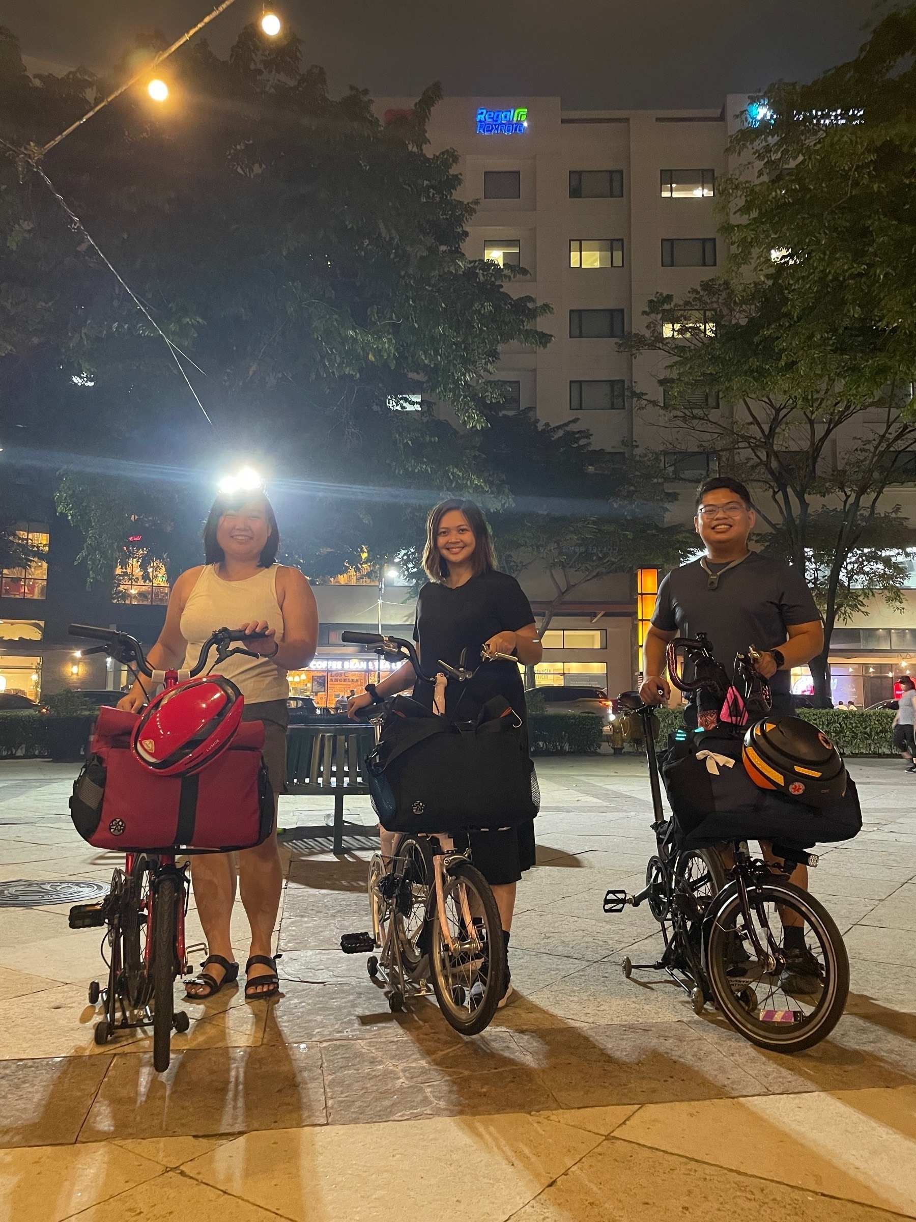 Chi posing with her two friends, Karen and Nix, next to their own trifold bikes and setups. On top of Chi's head, a street light is pointed towards the camera, so the top part of her head is obscured by the light.