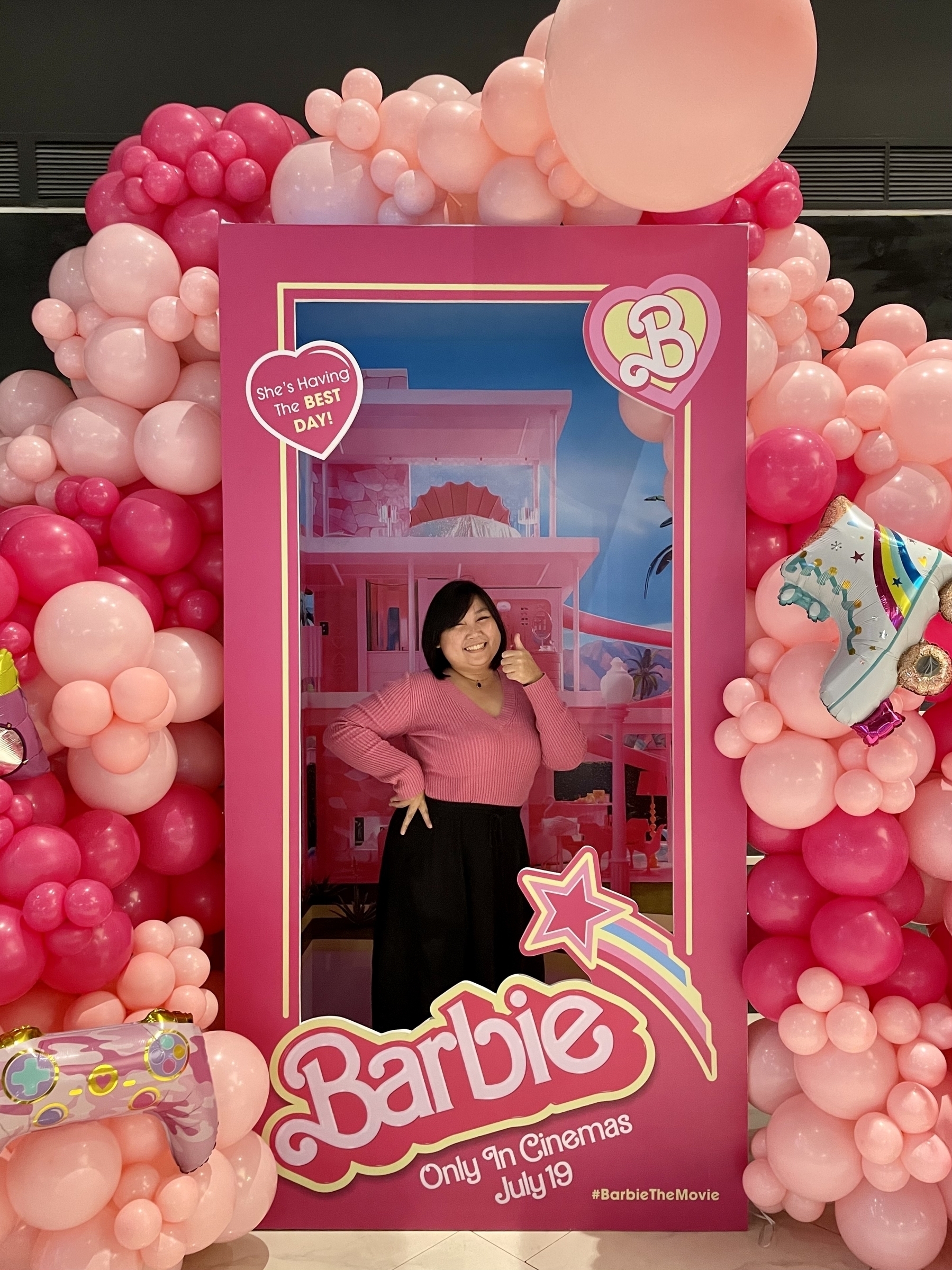 Chi posing in the Barbie Box promotional stand for the movie, dressed in a pink long-sleeved top and a black long skirt.