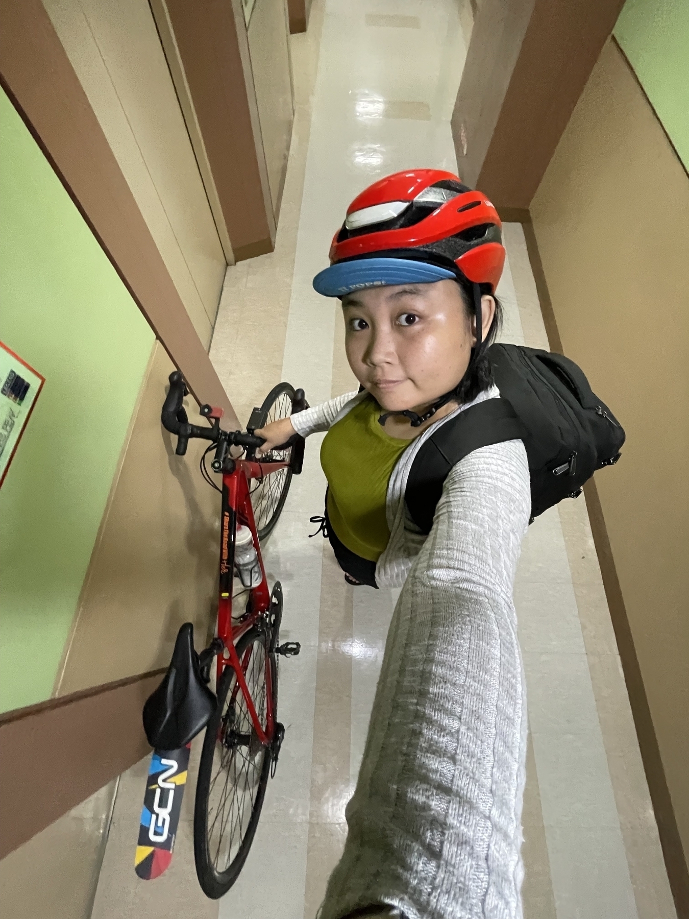 Wide angle selfie of Chi in her bike commute getup: she is wearing her normal, casual clothes, and has her backpack on which contains her Mac, her iPad, and other essentials. She is holding onto her red Merida road bike, which is also leaning towards the wall. She has her red Lumos helmet on, and a blue cycling cap.