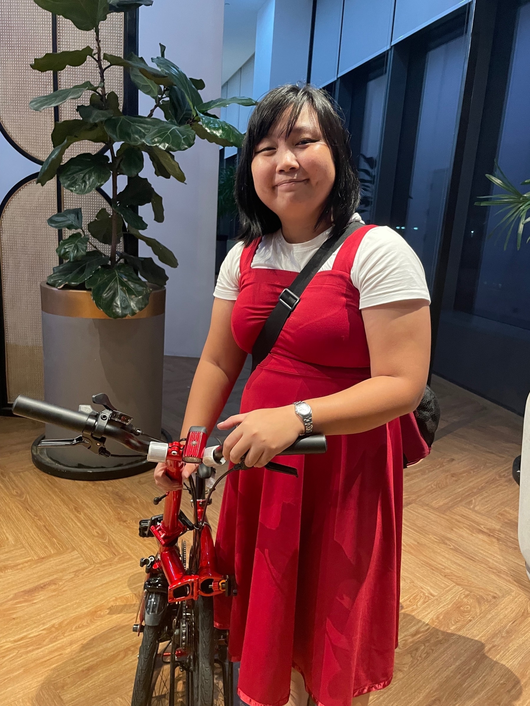 Chi posing with her red folded trifold bike. She is wearing a red long dress with a white undershirt, and slung on her shoulder is her red front bike bag. She is at a shared office space, with some indoor plants seen behind her.