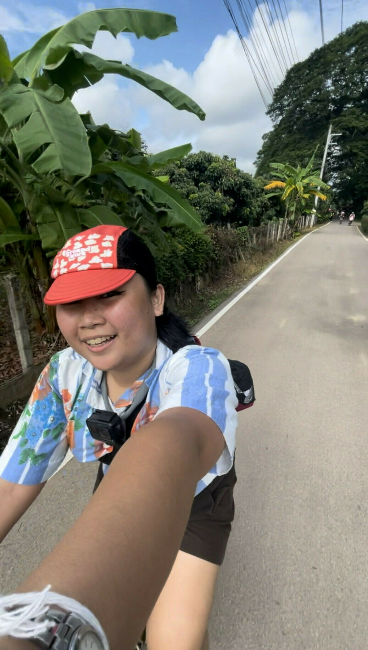 Wide angle selfie of Chi riding a bike, wearing her red popcorn cycling cap, kumot crop top, and black shorts, riding along one of the roads around the MueangKaen municipality at Chiang Mai, Thailand. Behind her are some trees, and the road is narrow enough for one person on a bike to pass with ease.