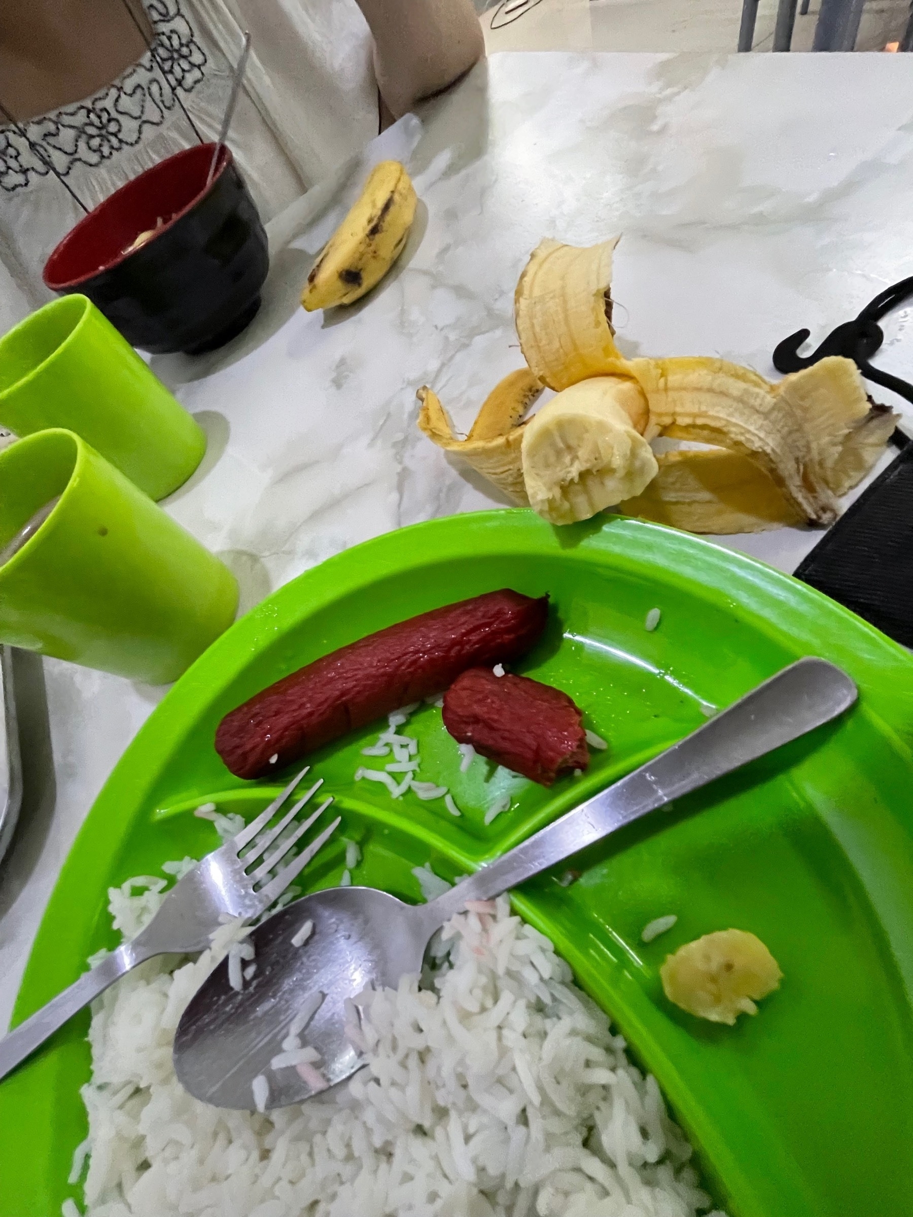 A green plate with rice, one hotdog, and a bitten-off banana are in view. Two green cups are also to the side, because Chi is eating breakfast with her mom.