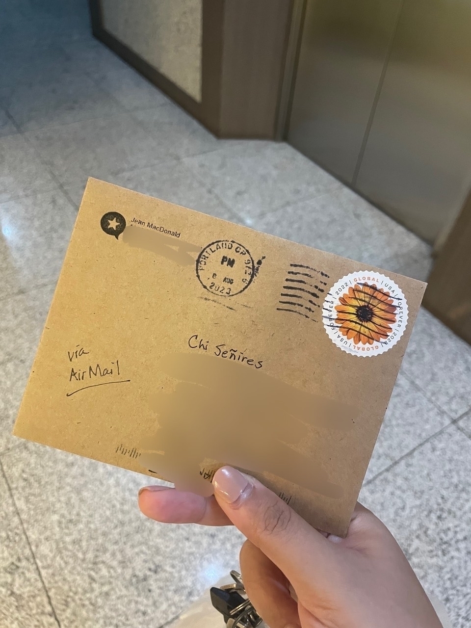 A brown envelope from Jean from Micro.blog, with address details obscured. There is a sunflower stamp on the upper right corner of the envelope, and a written underlined note on the lower left which reads, "via AirMail".