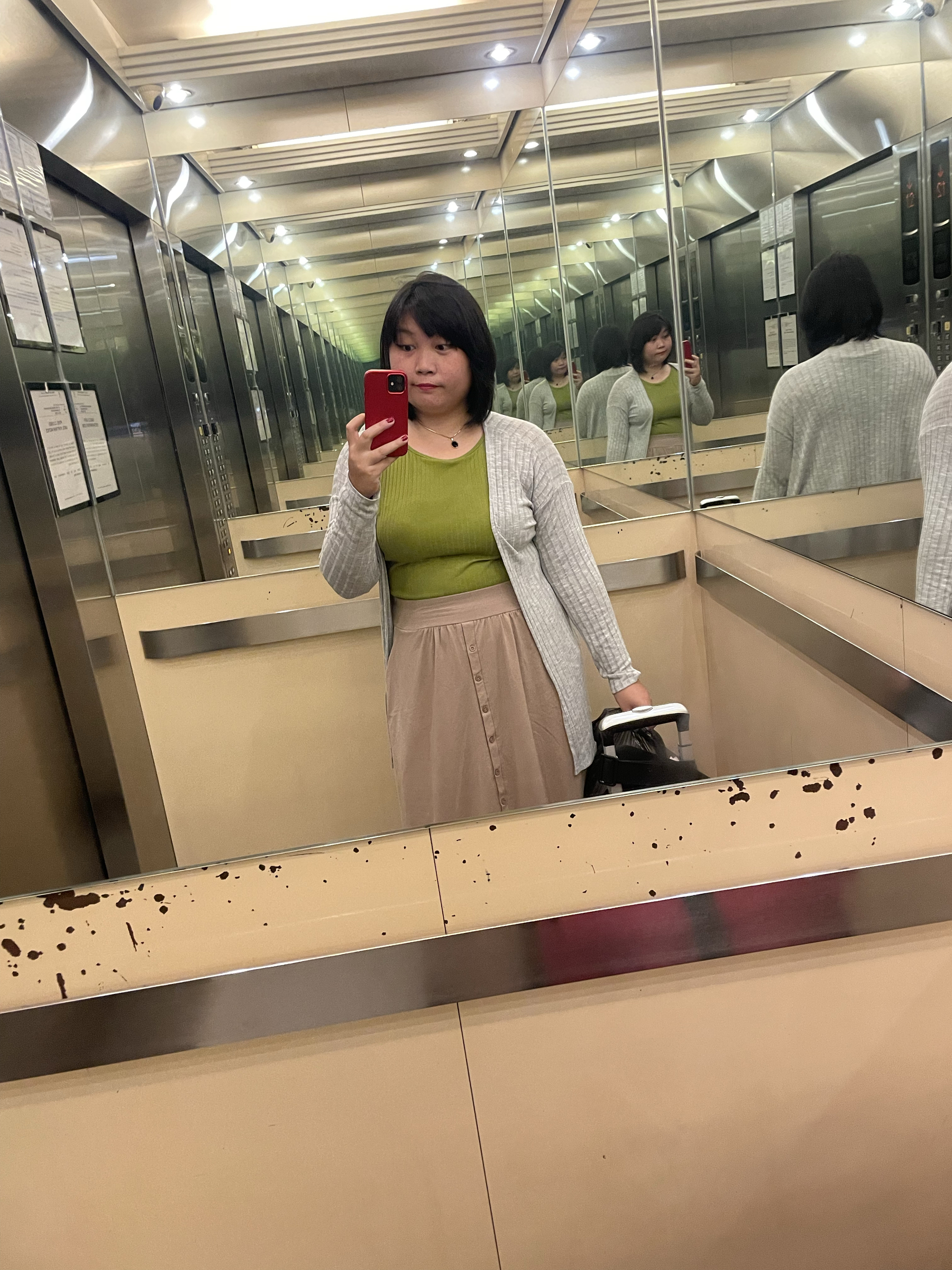 Elevator mirror selfie of Chi showing off her outfit: a green knitted sleeveless top and beige long A-line skirt, and a gray cardigan to complete the look. Seen peeking on the lower right portion of the mirror is her luggage.