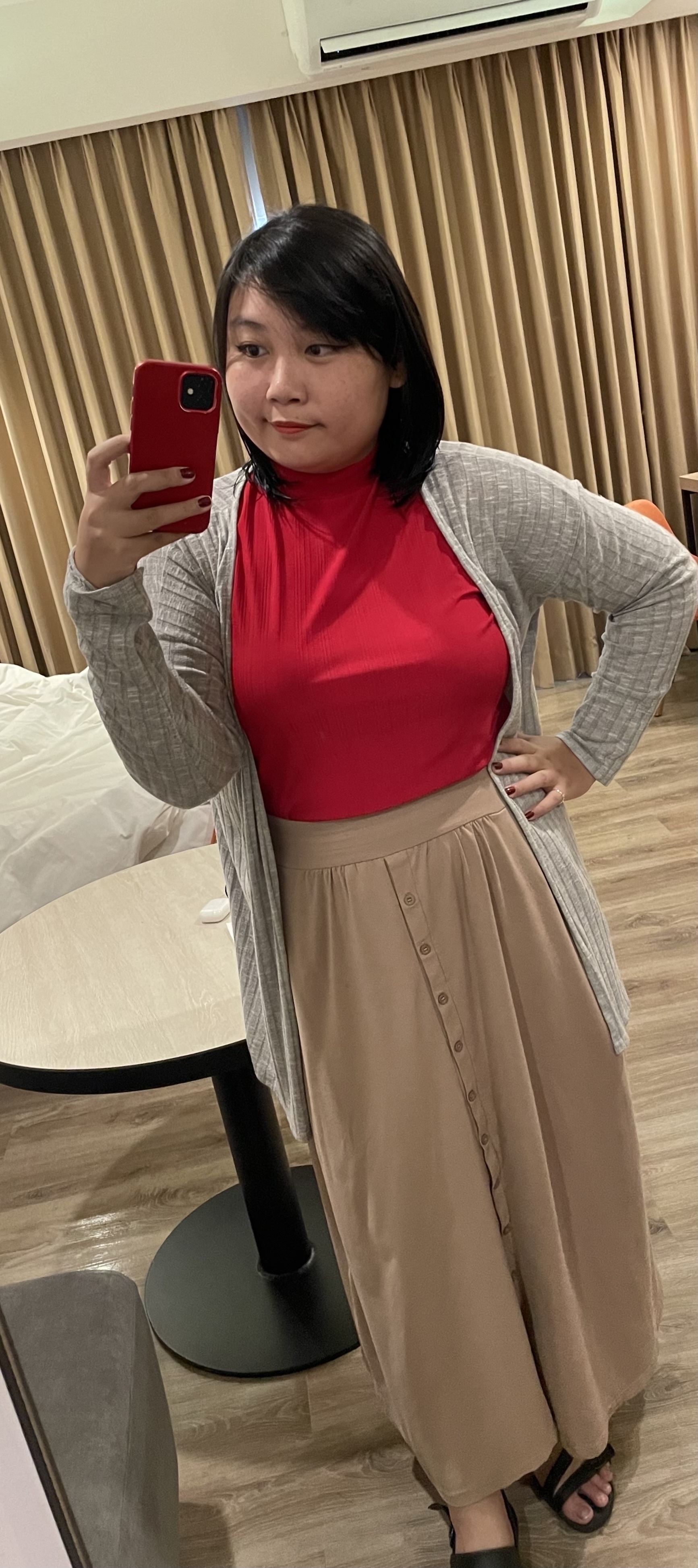 Chi's ouftit, which is a red turtleneck sheer top tucked in her long beige skirt, and with a gray cardigan and black sandals. She's also wearing her signature makeup look, which is cat eye eyeliner and red lipstick.