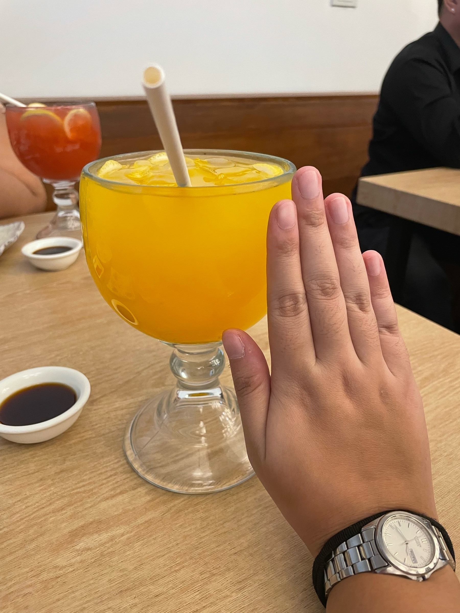 Chi's glass filled with mango juice, with a really big glass. The size is as big and wide as her hand, which is positioned right next to the glass for comparison. In the background, her companion's drink is also visible: a red drink, but Chi forgot what it was.