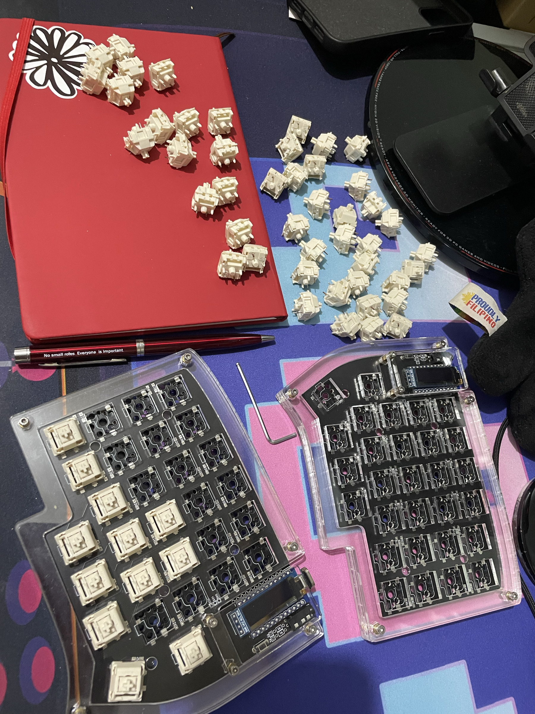 photo of Chi’s desk with a bunch of keyboard switches scattered on top of notebook, and some keyboard switches also inserted already in the split keyboard frame. The printed circuit board can be seen through the split keyboard’s acrylic case.