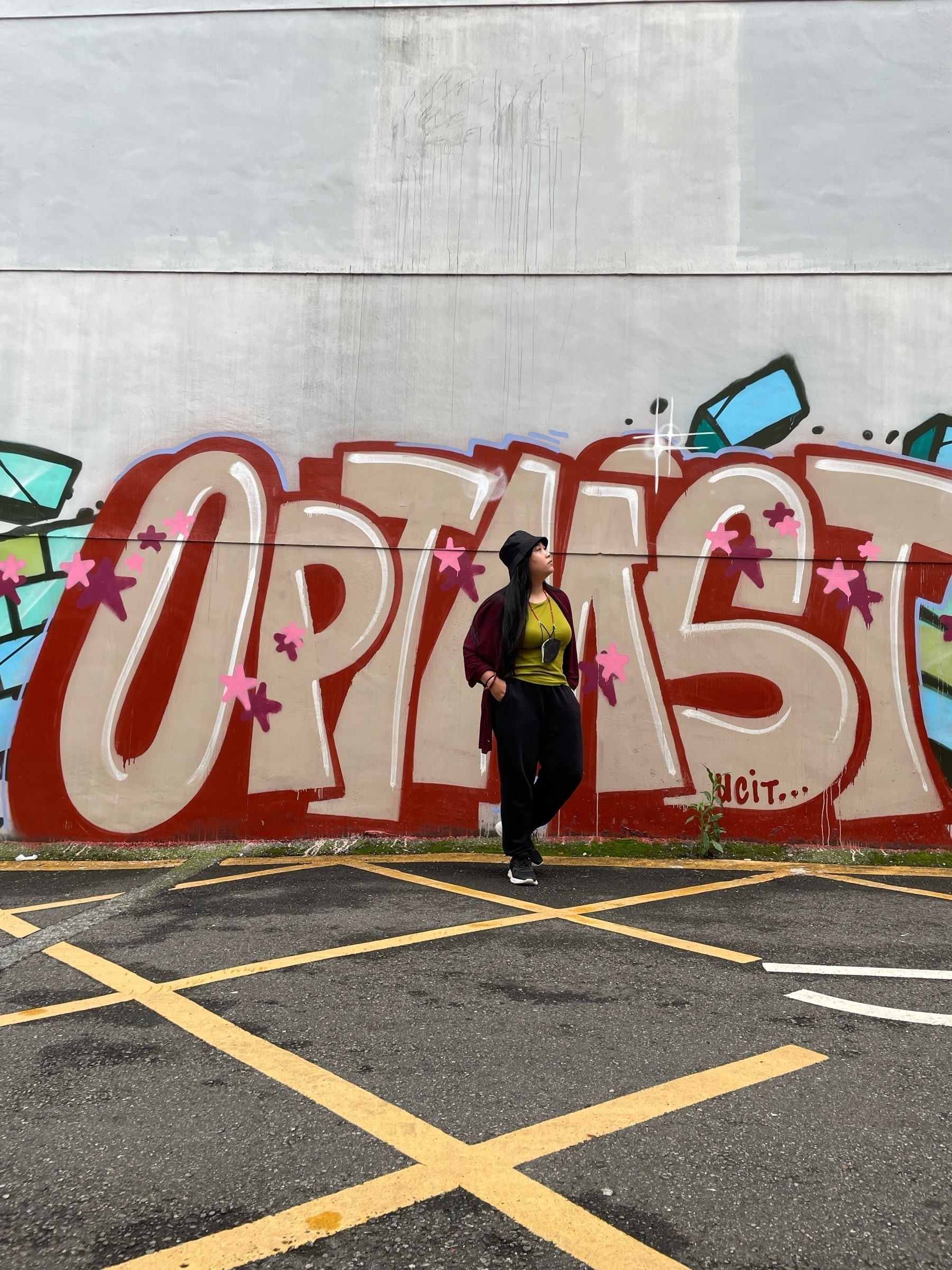Chi in front of a gray wall at a parking lot area with the graffiti of the word OPTIMIST drawn with a red-maroon outline and small pink stars. Chi is wearing a black bucket hat, a green knitted top, black jogging pants, and black sneakers. Around her neck is a black KN95 mask tied to a red necklace. She has a red shawl repurposed to be like a cardigan over her shoulders. She is looking away from the photo, with one leg propped behind her, and her hands are in her pockets, suggesting that she is deep in thought.