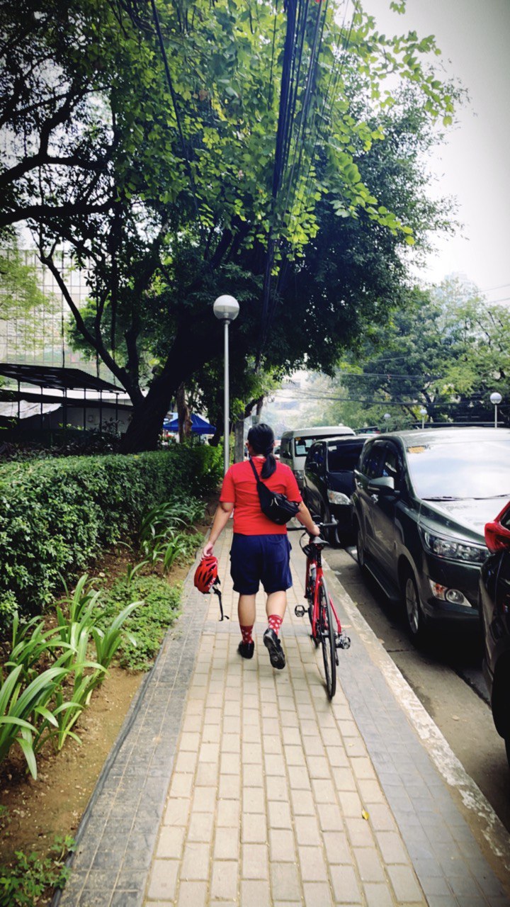 Photo of Chi walking with her road bike on the sidewalk. The point-of-view is from her back.