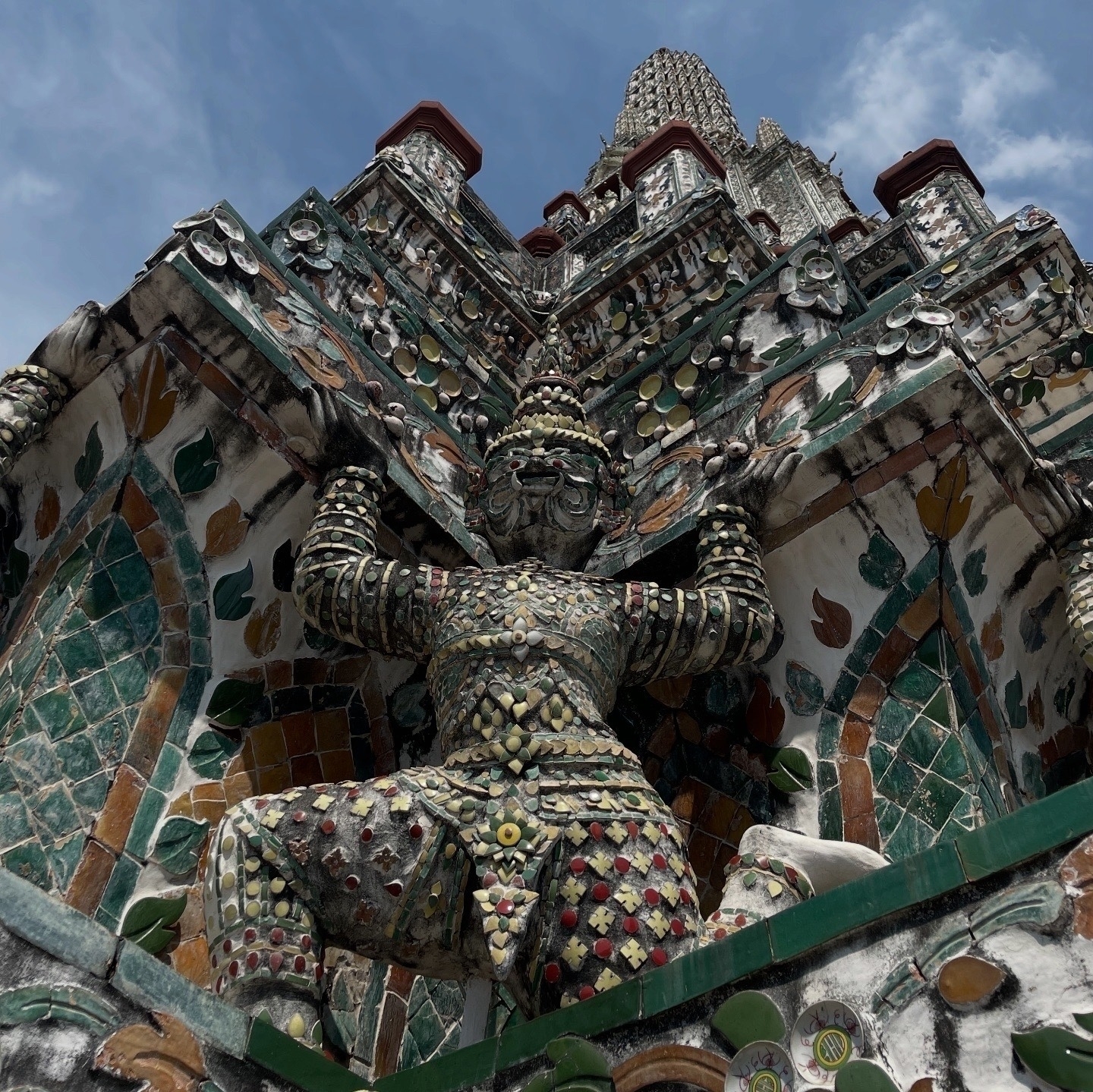 Close-up view of a giant demon yaksha guardian figure at Wat Arun, shot from below. The figure is decorated with intricate ceramic mosaic tiles in various colors, standing against a background of floral mosaic patterns in green, white, and gold. The ornate details of the temple's spire can be seen rising behind the yaksha against a blue sky. The low angle emphasizes the imposing nature of the guardian statue and showcases the detailed ceramic work.