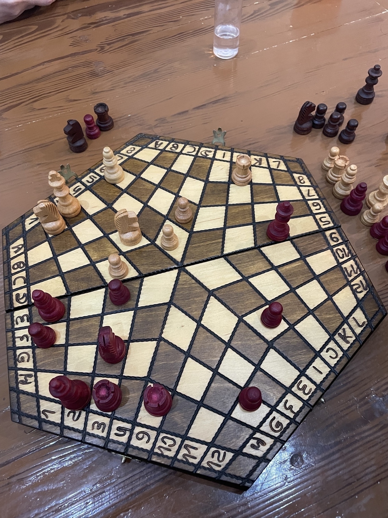 Late-game position in a three-player chess match on a hexagonal board with numbered edges. Only red and light wooden pieces remain on the board, as the dark wooden side has been eliminated. Captured pieces are placed off the board, with a water bottle visible in the background.