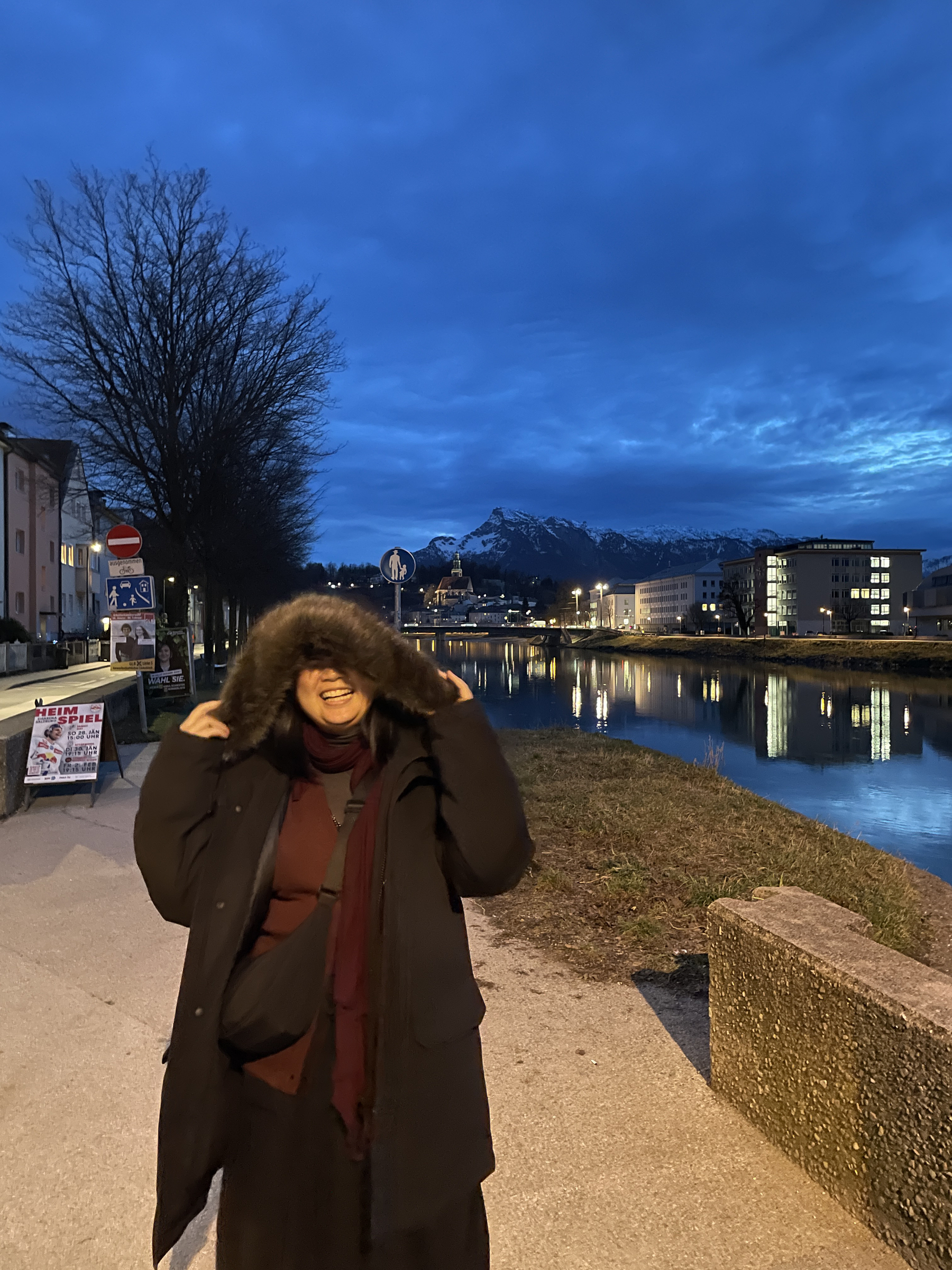Candid photo of Chi next to the Salzach River wearing her boyfriend’s big winter jacket, with the hood going on top of her head and seemingly eating her because of how big the jacket is. The sky is a dark blue, with more clouds in sight, and the sun has set already.