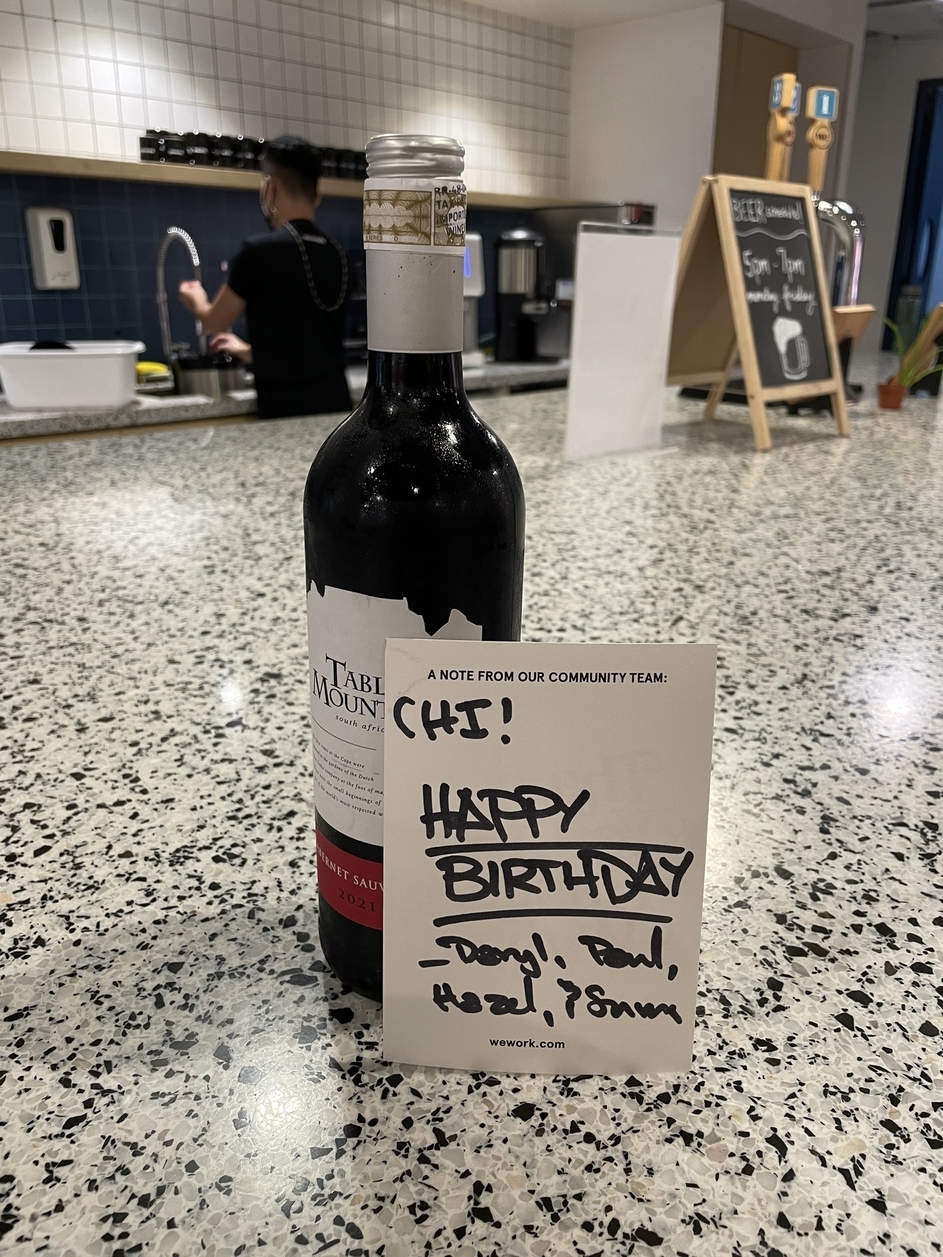 A wine bottle is on a marbled countertop with a greeting card in front of it. The text reads, "A note from our community team: Chi! Happy birthday Daryl, Paul, Hazel, & Sumana"