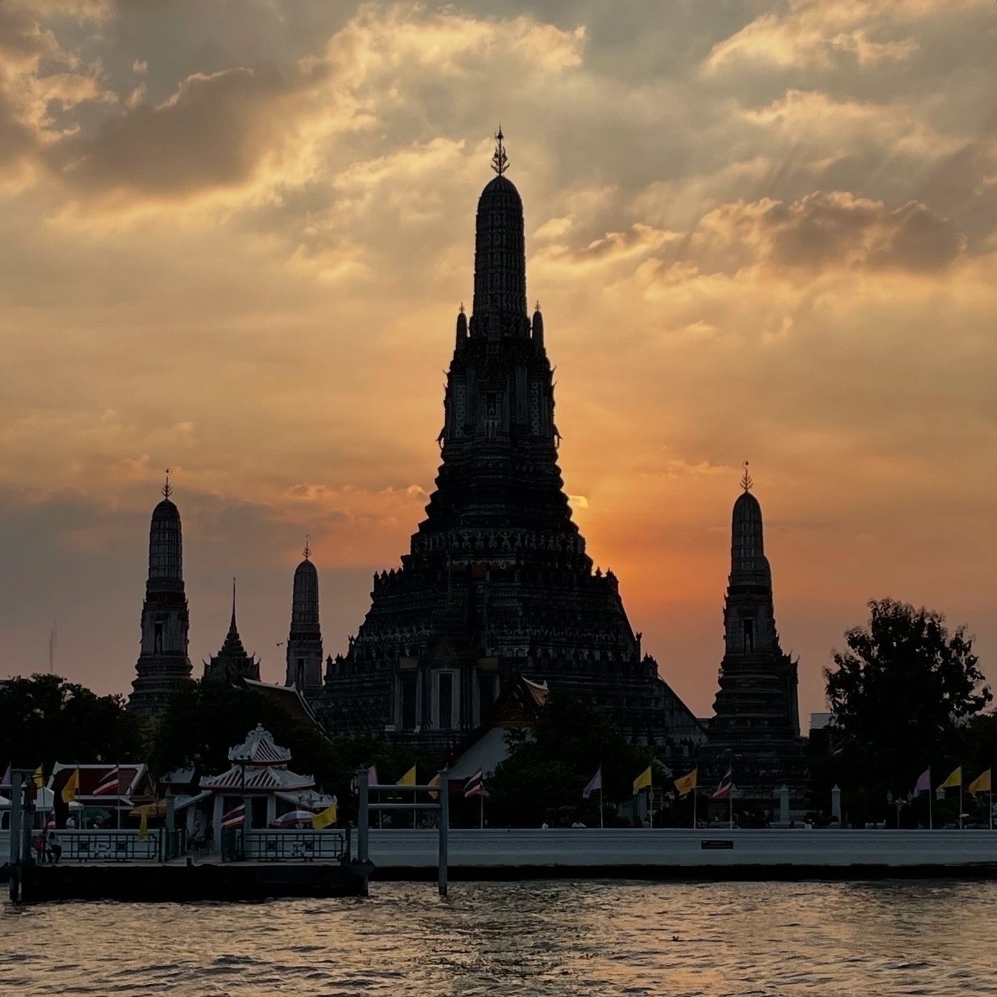 Sunset view of Wat Arun from across the Chao Phraya River. The temple's iconic prangs are silhouetted against a dramatic orange and golden sky with scattered clouds. The river's choppy waters reflect the sunset's warm colors, creating golden ripples in the foreground. A pier with Thai and colored flags can be seen along the riverbank. This view captures why the temple is known as the Temple of Dawn, though photographed at dusk.