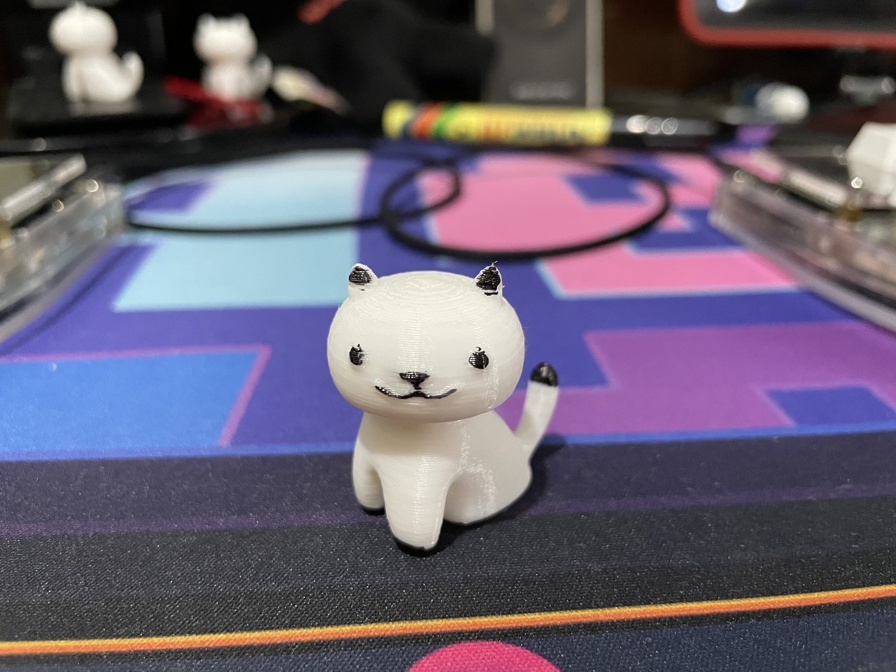 A 3d-printed white cat with distinctive features like its ears, eyes, nose, mouth, paws, and tail tip colored in with a black marker. The cat is seated on a computer deskmat.