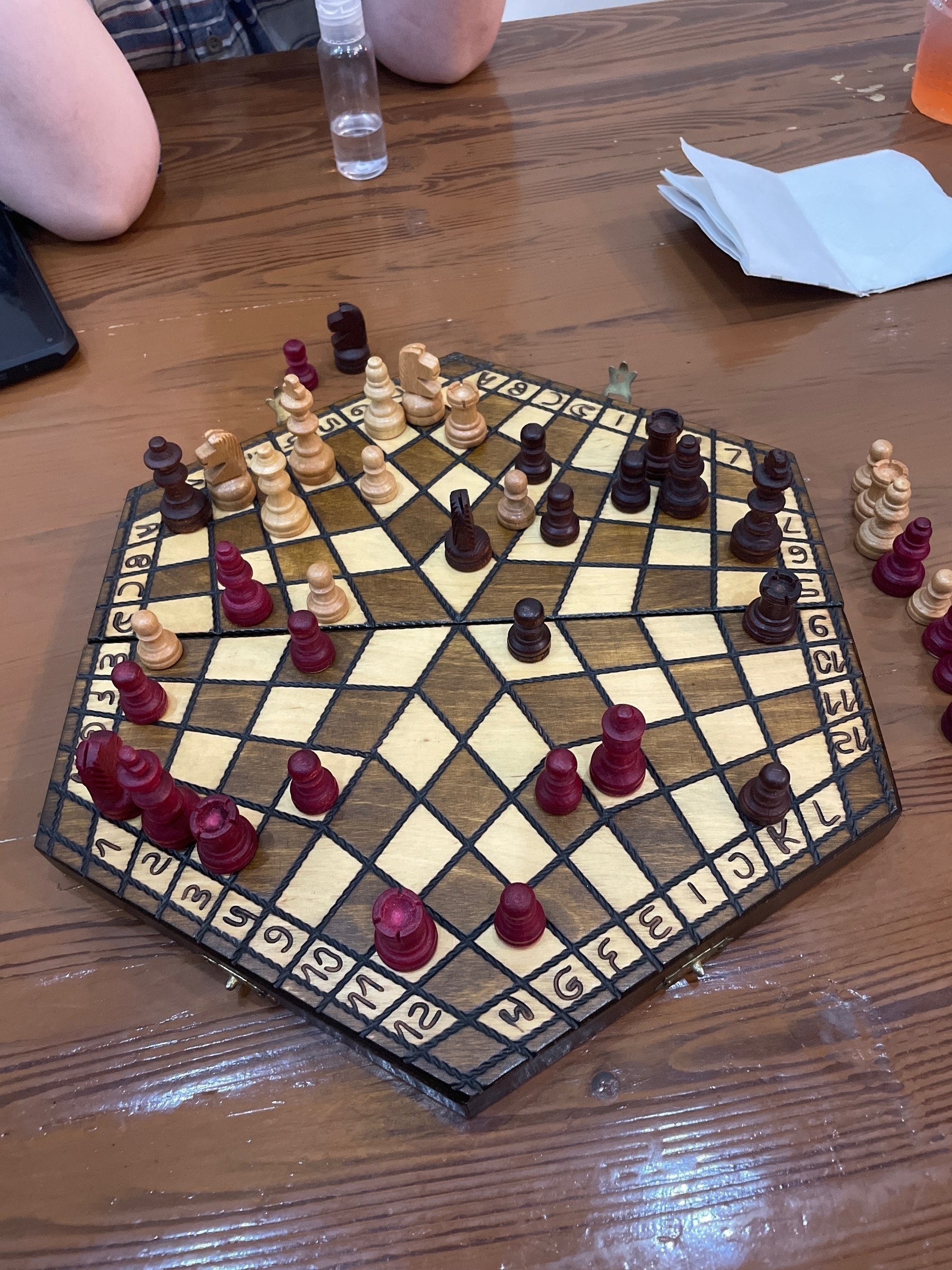 A three-player chess game in progress on a hexagonal board with alternating light and dark squares. The board features pieces in three colors—red, light wood, and dark wood—with some pieces captured and placed aside on the wooden table.