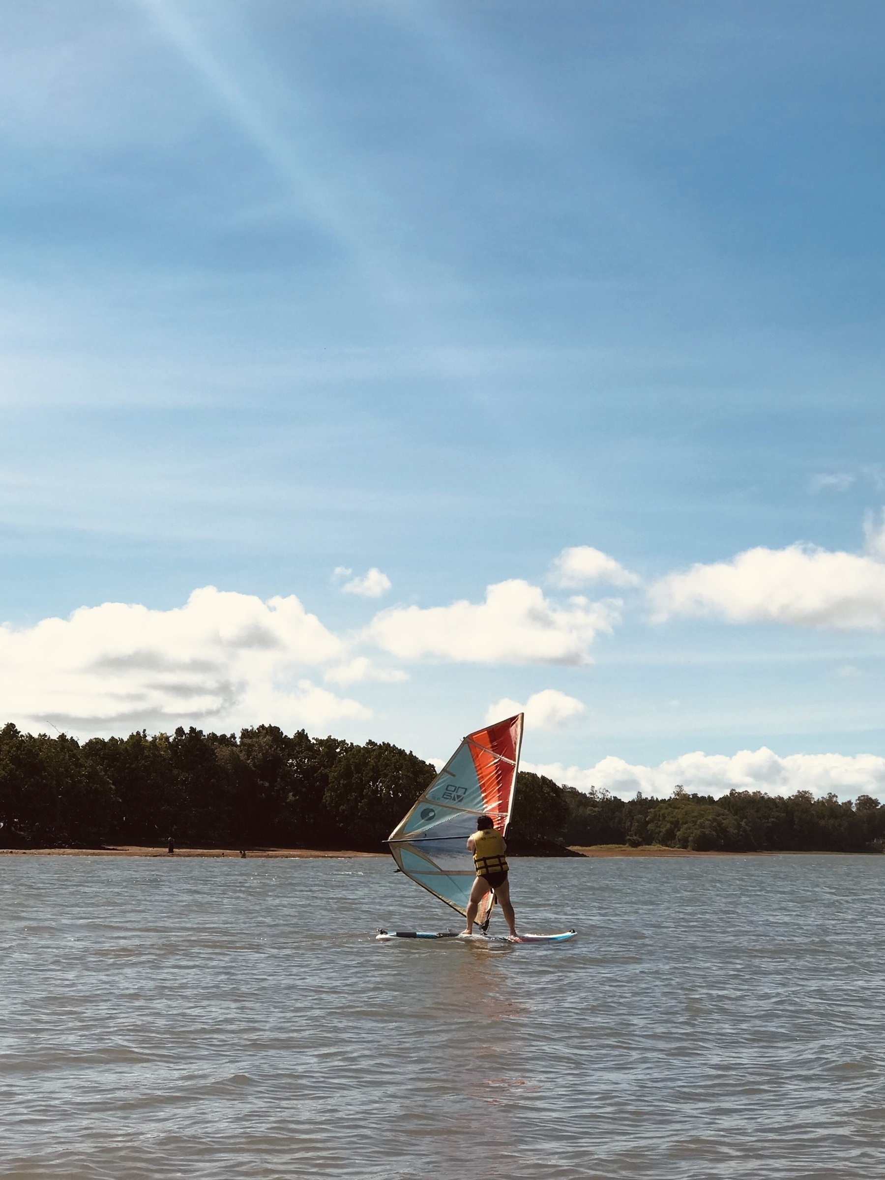 Chi is controlling a windsurfer going to the right of the point-of-view while in the middle of Caliraya Lake in Laguna. The blue sky takes up majority of the shot, with a few clouds and some sun rays visible. In the background, a forest can be seen lining up right before the shore that draws a line in the middle of the shot.