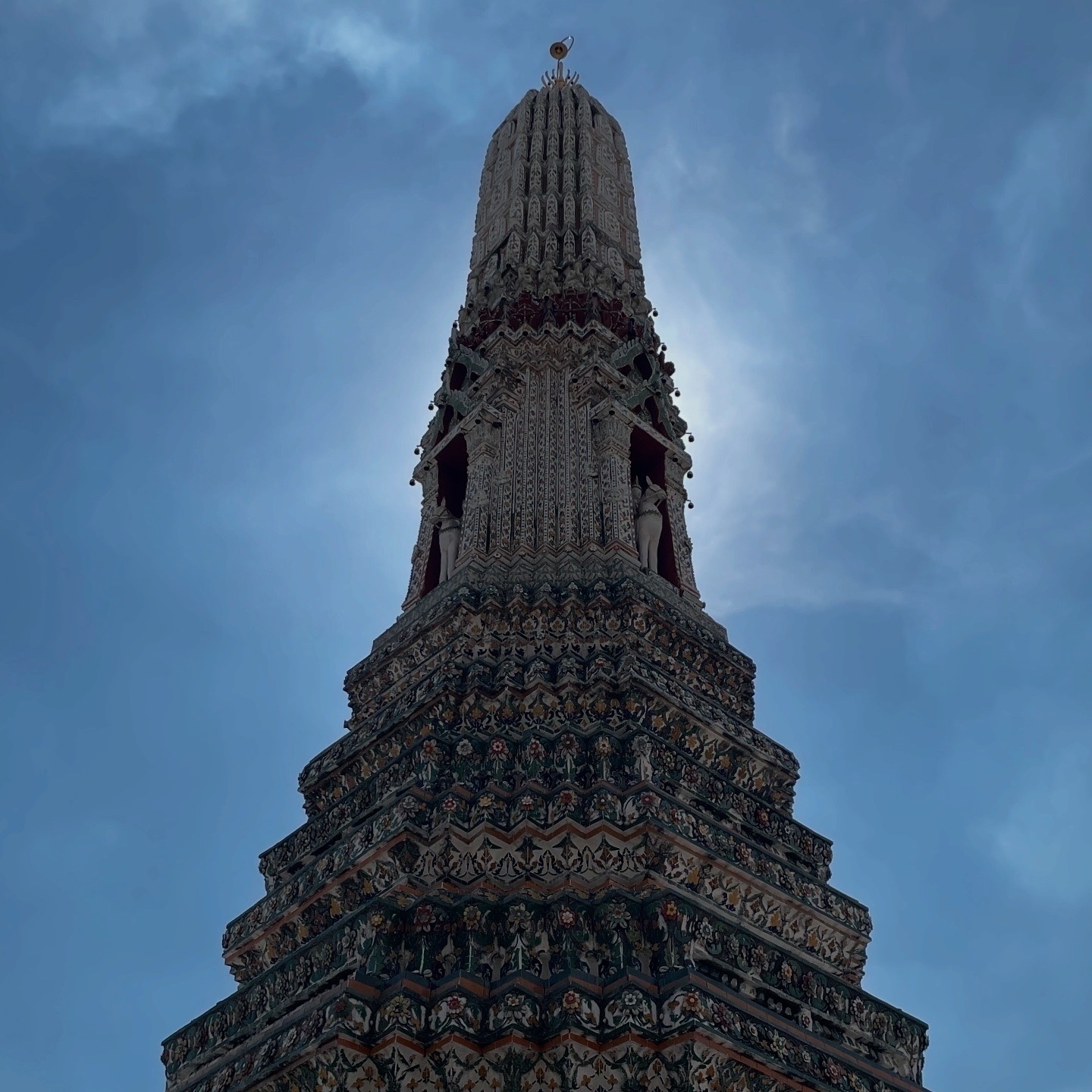 A dramatic upward view of one of Wat Arun's iconic prangs (spires), showing its detailed Khmer-style architecture. The prang is decorated with intricate ceramic tiles and ornamental stuccoes in layers, with demon yaksha figures visible at its base. The spire rises against a blue sky with wispy clouds, creating a striking silhouette. Shot from below, the perspective emphasizes the tower's impressive height and detailed ornamentation.