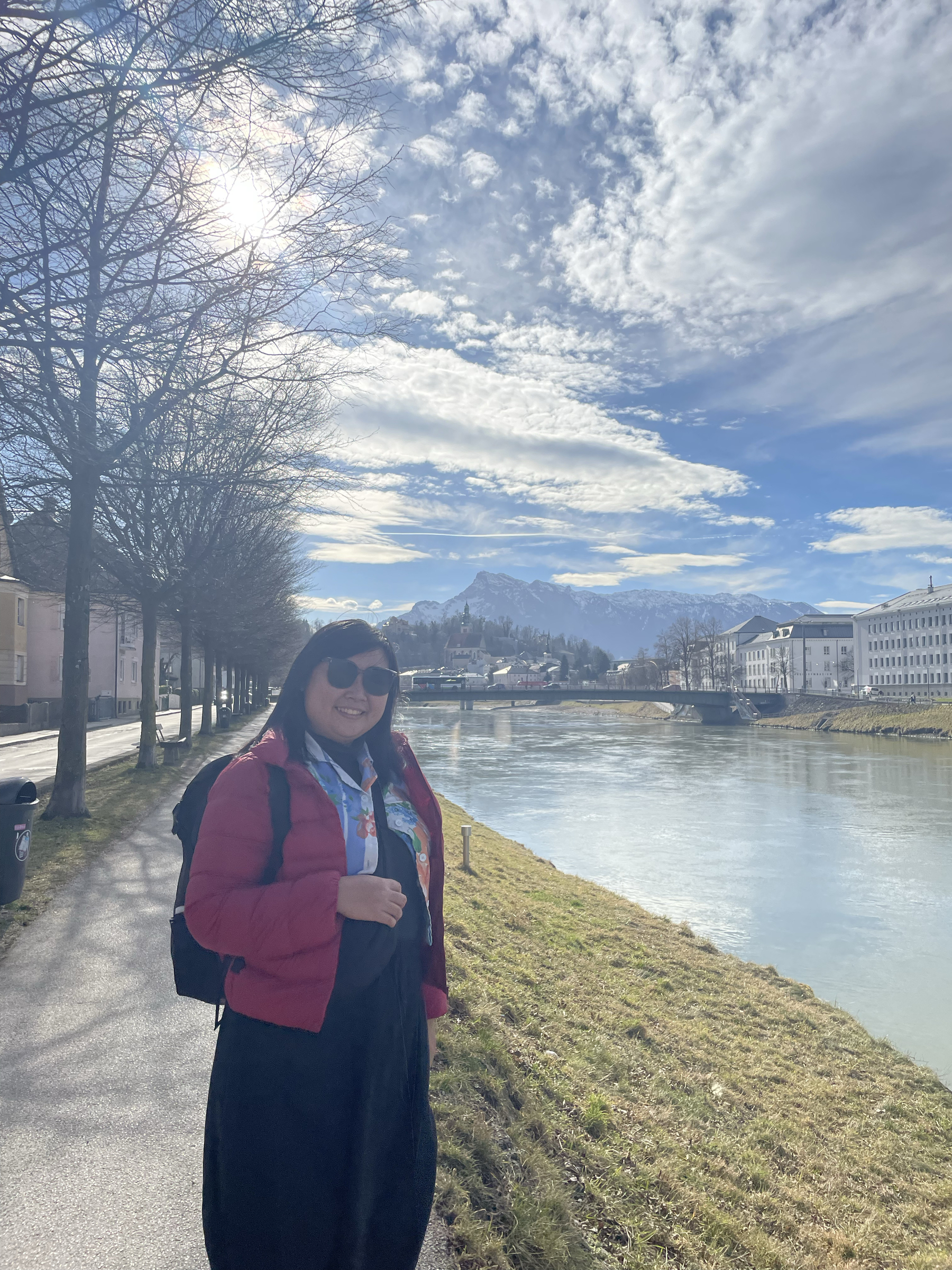 Photo of Chi next to Salzach River. It is a sunny day, with some clouds seen but not blocking the sunlight. Chi is wearing shades, and a red puffer jacket hiding her outfit of a blue crop top that has the pattern of an old blanket from her childhood, a black turtleneck, and a black long skirt. She has her backpack behind her back. A row of leaf-less trees are seen behind her.
