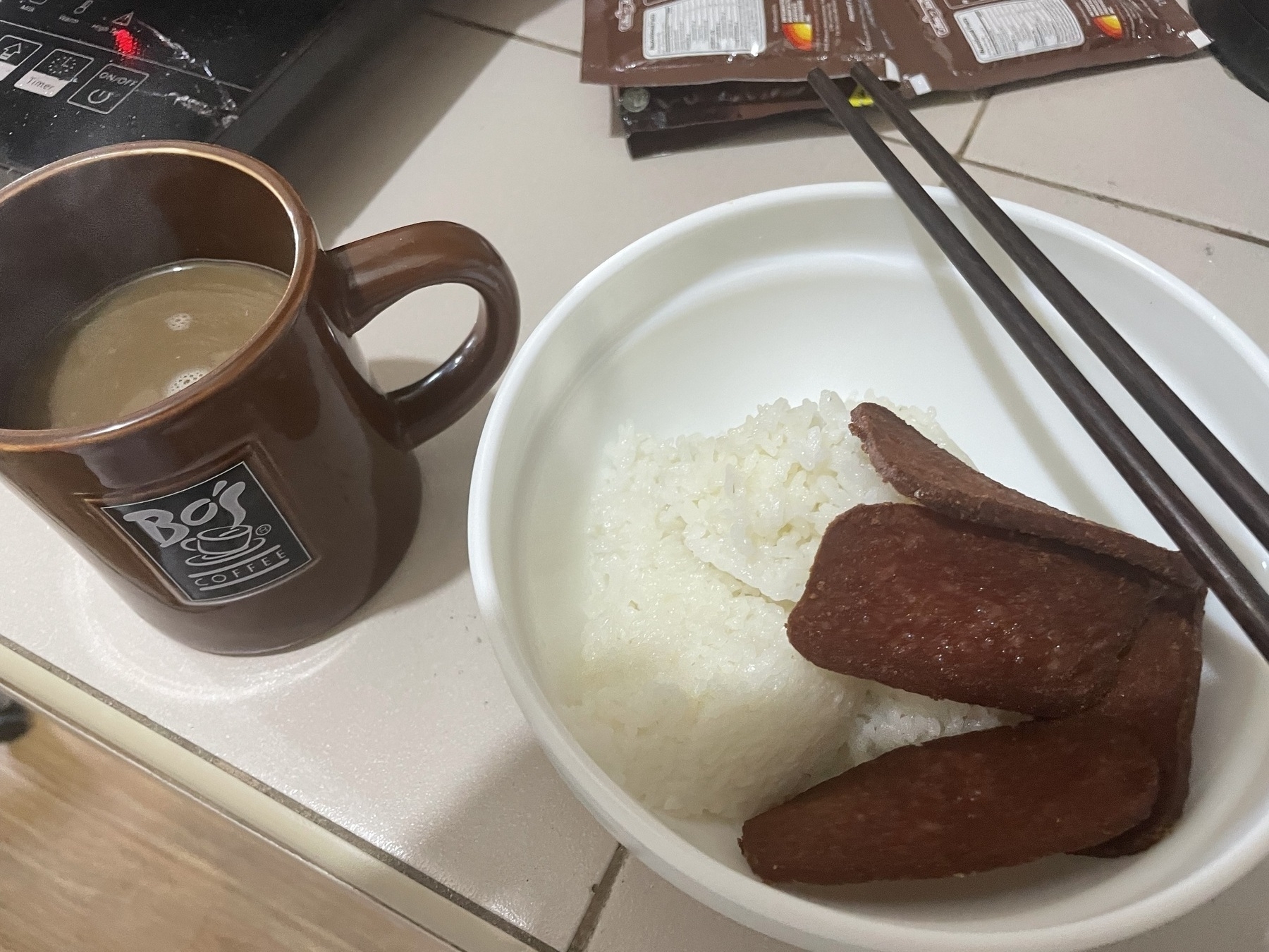 Chi's usual breakfast meal, which is a hot coffee with sugar and creamer and fried luncheon meat with a couple of scoops of rice. Her utensils are chopsticks, and her meal is placed in a bowl.