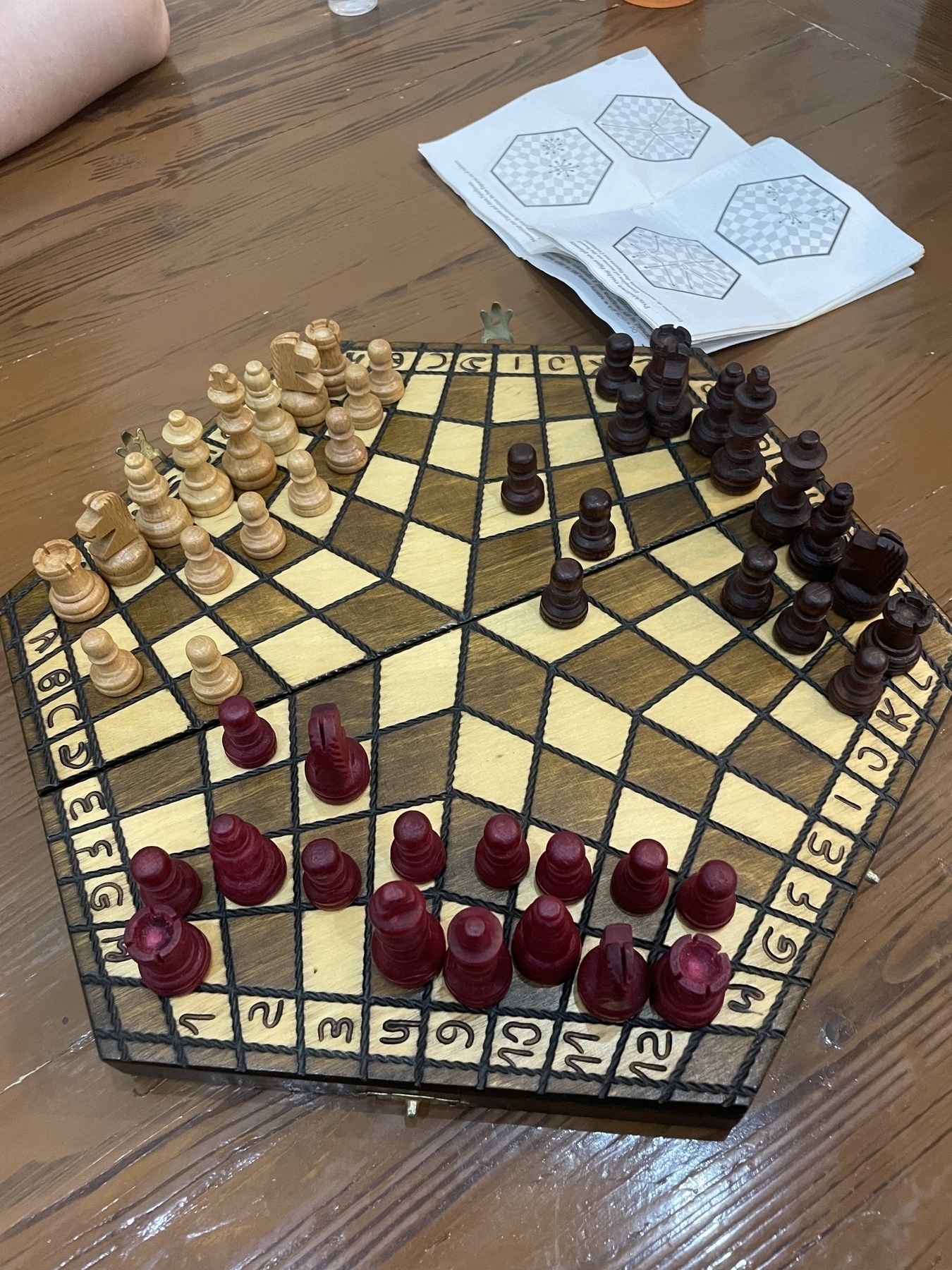 A hexagonal three-player chess board with dark and light squares, showing chess pieces arranged during gameplay. The board has numbered edges and contains light wooden, dark wooden, and red pieces. The instructions sheet of paper with hexagonal diagrams is visible in the background.