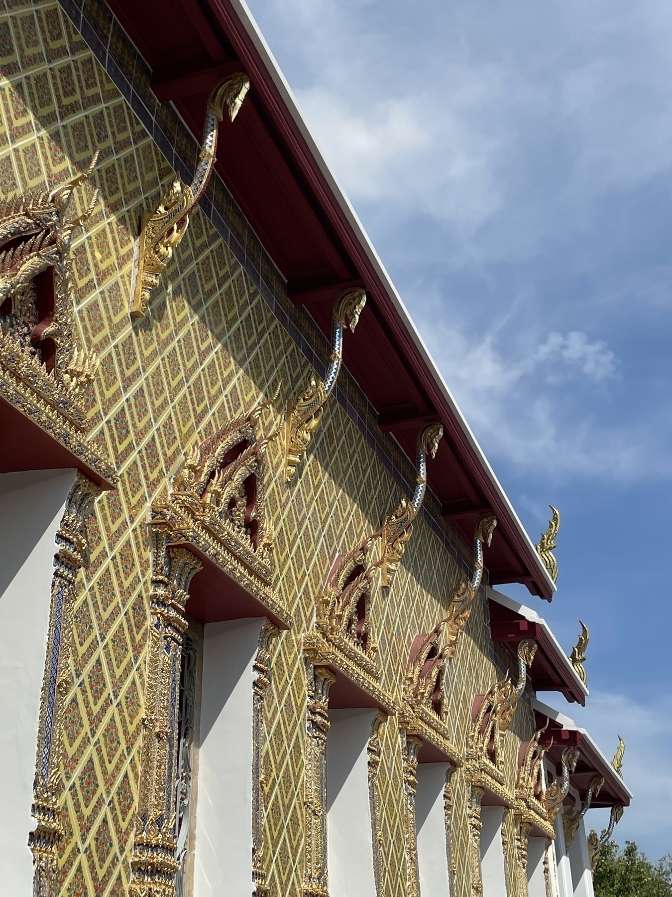 Ornate exterior detail of a Thai Buddhist temple, showing a row of gilded columns decorated with intricate traditional Thai patterns. The columns feature gold-leafed serpent (naga) designs and diamond-patterned mosaic work in gold and white against a red roof trim. The image is shot from a low angle against a bright blue sky.