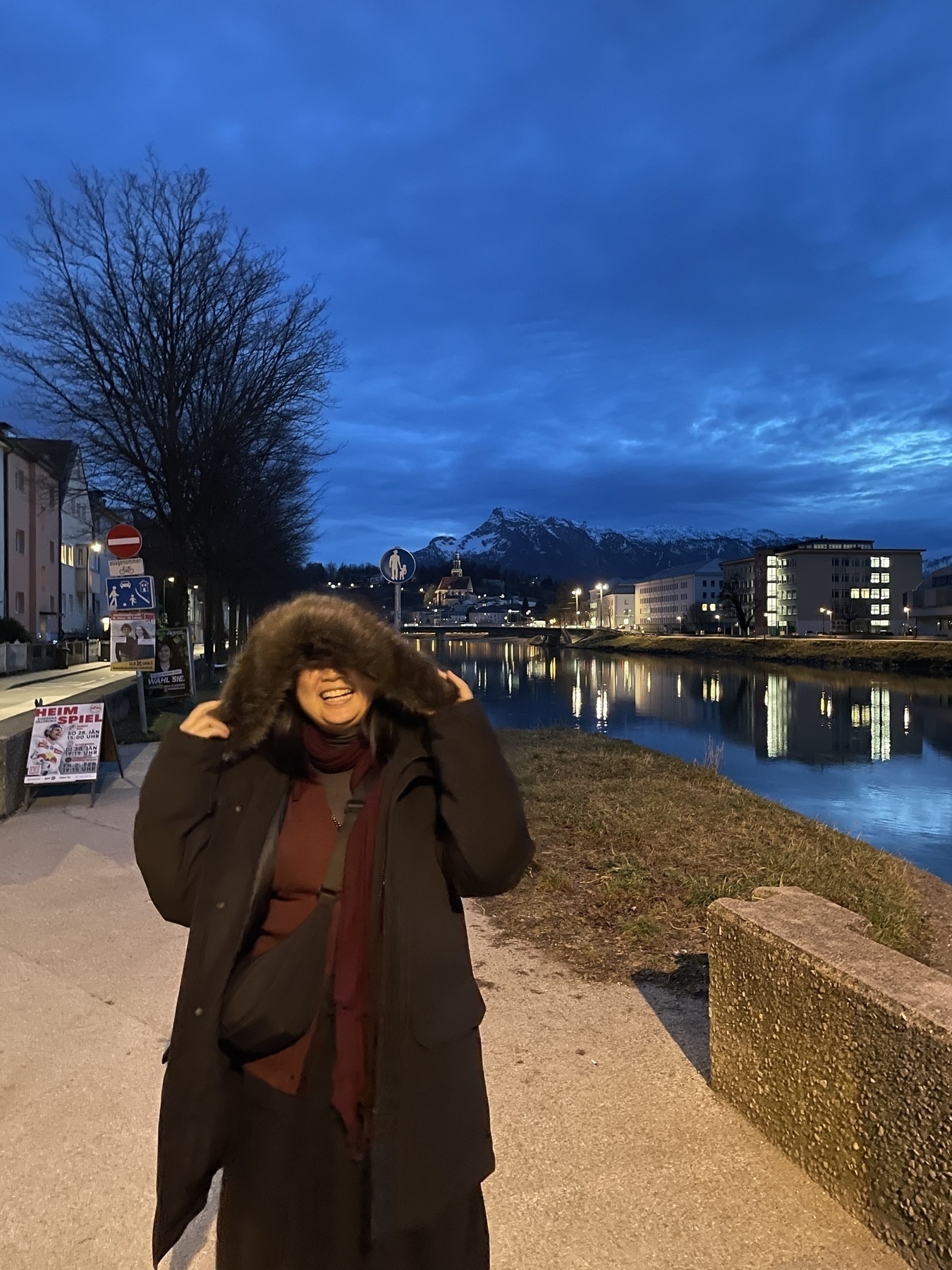 Chi is laughing while wearing her boyfriend's black big winter jacket with a fluffy hoodie, because it looks like she's being eaten by the jacket with the hoodie on top of her head. Behind her is the Salzach in Salzburg, Austria. The sky is dim, because it's early evening.