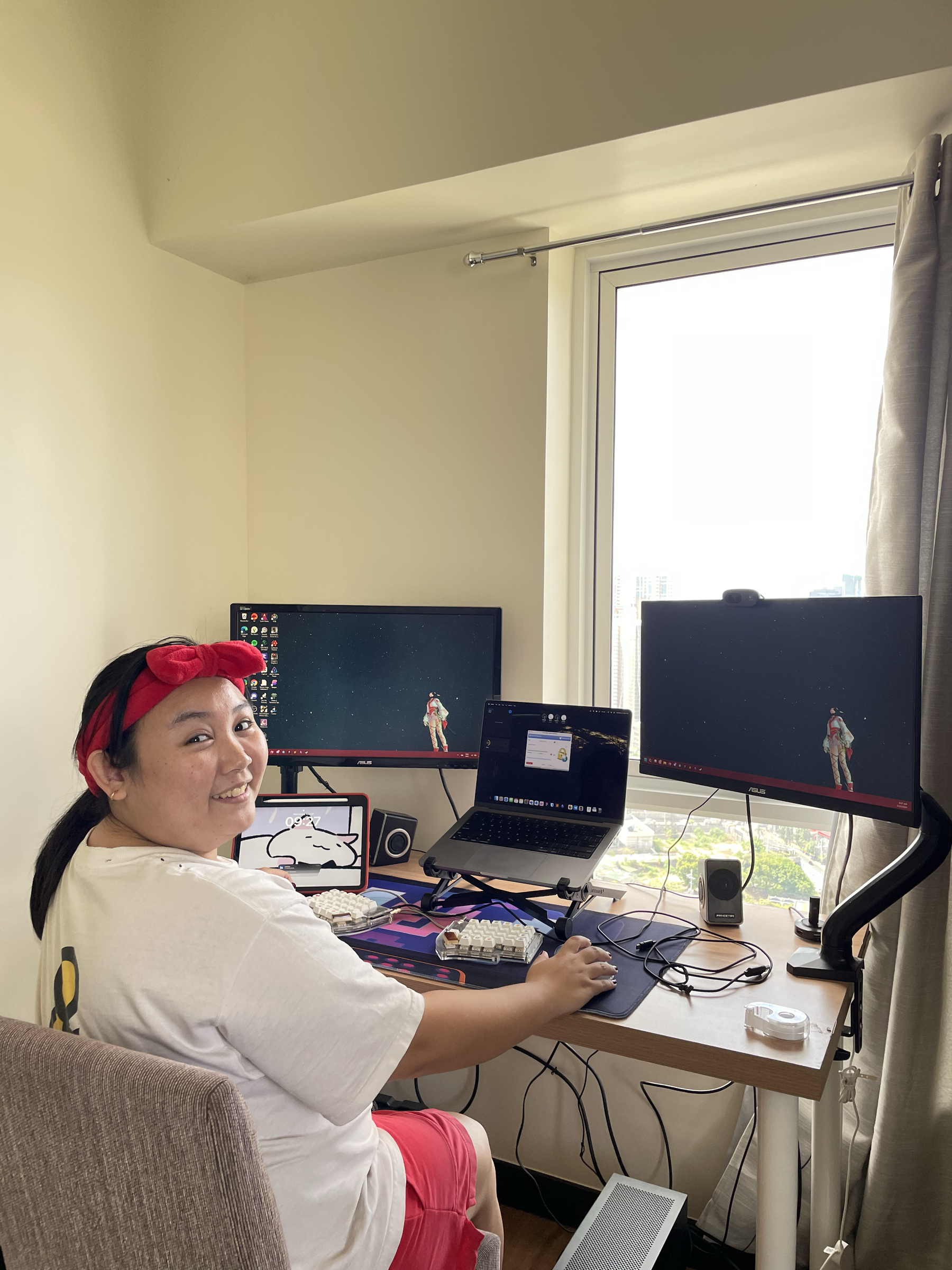 Chi looking at the camera and smiling while at her home workstation, consisting of two monitors mounted on monitor arms, an iPad propped up, and a Macbook Pro elevated with a laptop stand in the middle of her two monitors. On her desk is her split keyboard, her trackball mouse which Chi is holding, and a full deskmat. The chair Chi is sitting on is still a normal dining chair, not her computer chair. Her setup is facing the window, letting natural light in.
