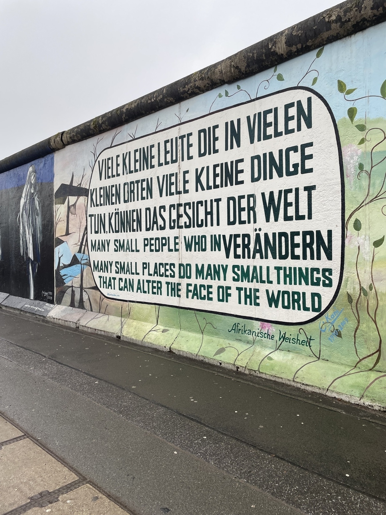 Urban art found on the East Side Gallery with text that reads, first in German: “Viele kleine Leute die in vielen kleinen Orten viele kleine Dinge tun, können das Gesicht der Welt verändern.” and then in English: “Many small people who in many small places do many small things that can alter the face of the world.” underneath the text block is the credits to "Afrikanische Weisheit"—African wisdom in english.