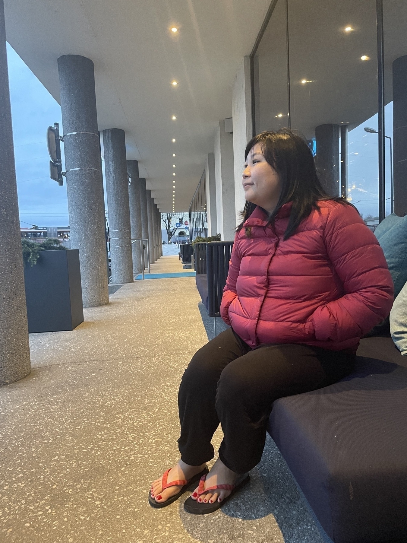 Chi is seated on a sofa outside of her hotel and is looking towards the left of the photo, which is where the road is but it's not seen. She's wearing a red puffer jacket and black leggings to keep warm, but also is seen wearing slippers.