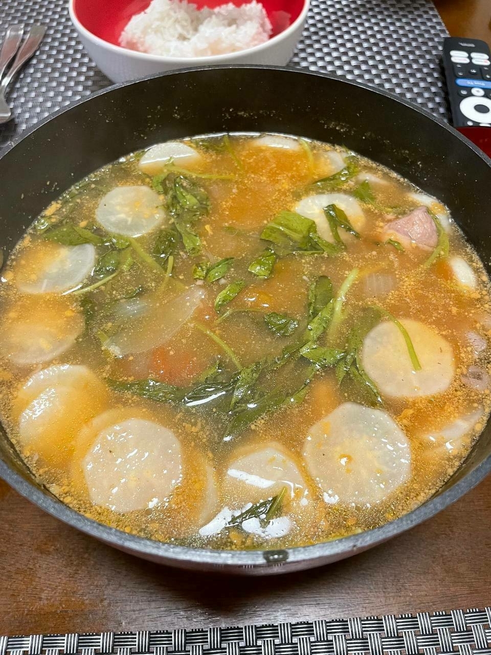 Sinigang na baboy in a pot and placed on a dining table. A bowl of rice is seen behind the pot, as well as some utensils and a placemat. Pieces of labanos and kangkong are floating on the top of the sabaw, and the sinigang looks like its signature orangey color.