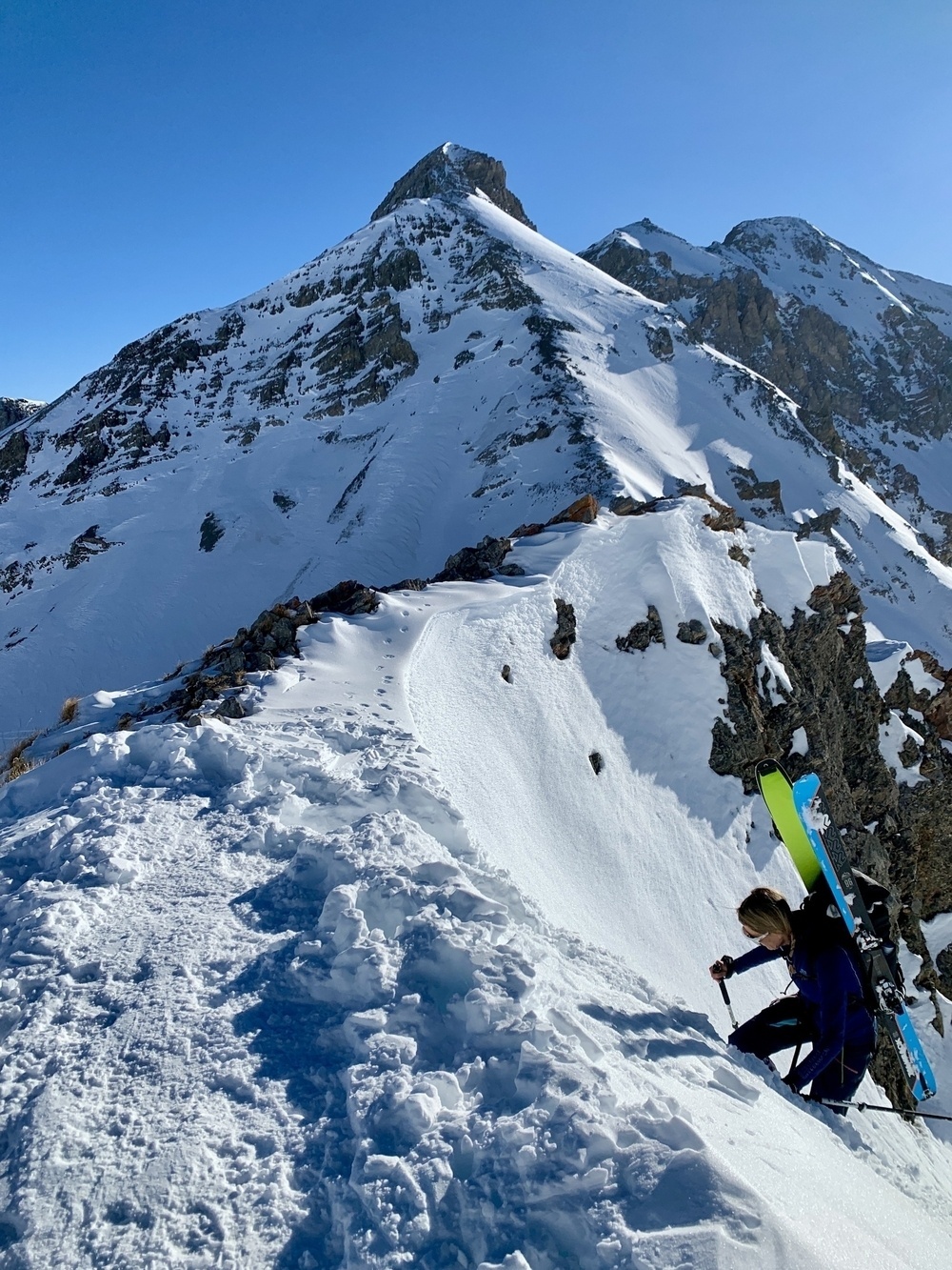 A person is climbing a snow-covered mountain carrying skis, with another peak in the background under a clear blue sky.