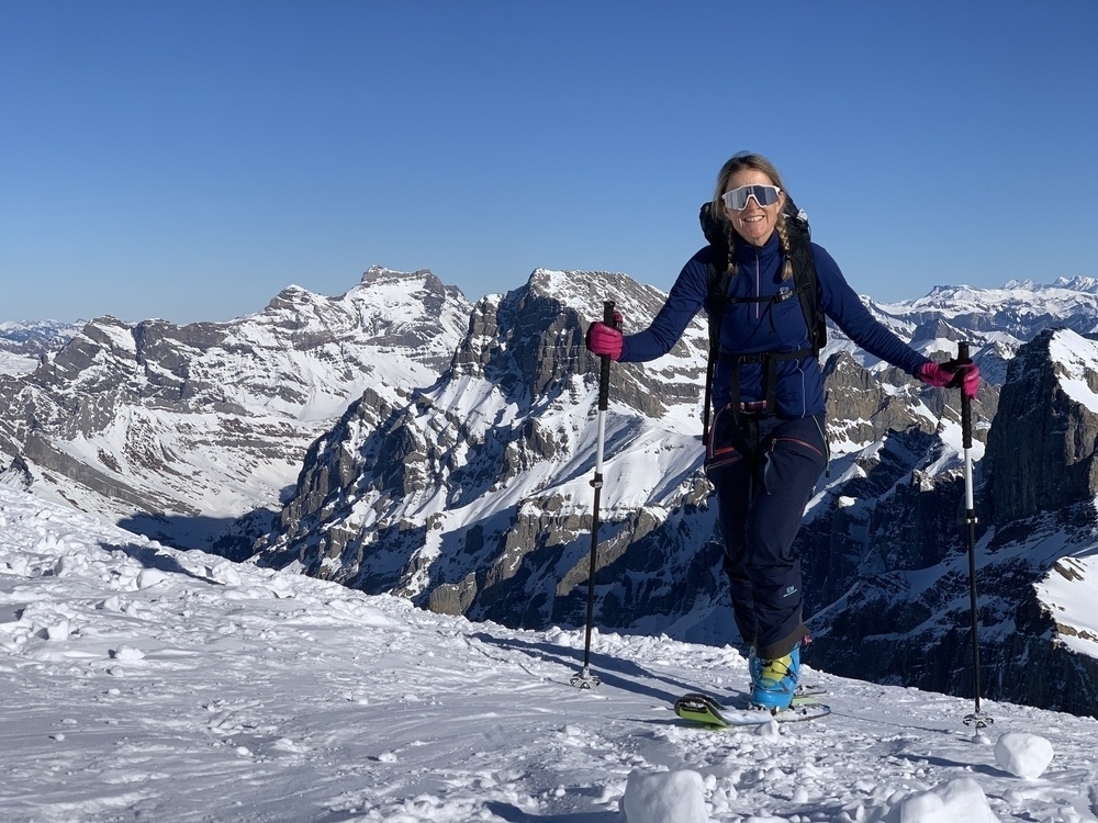 A person is skiing on a snowy mountain with a clear blue sky and rugged peaks in the background.