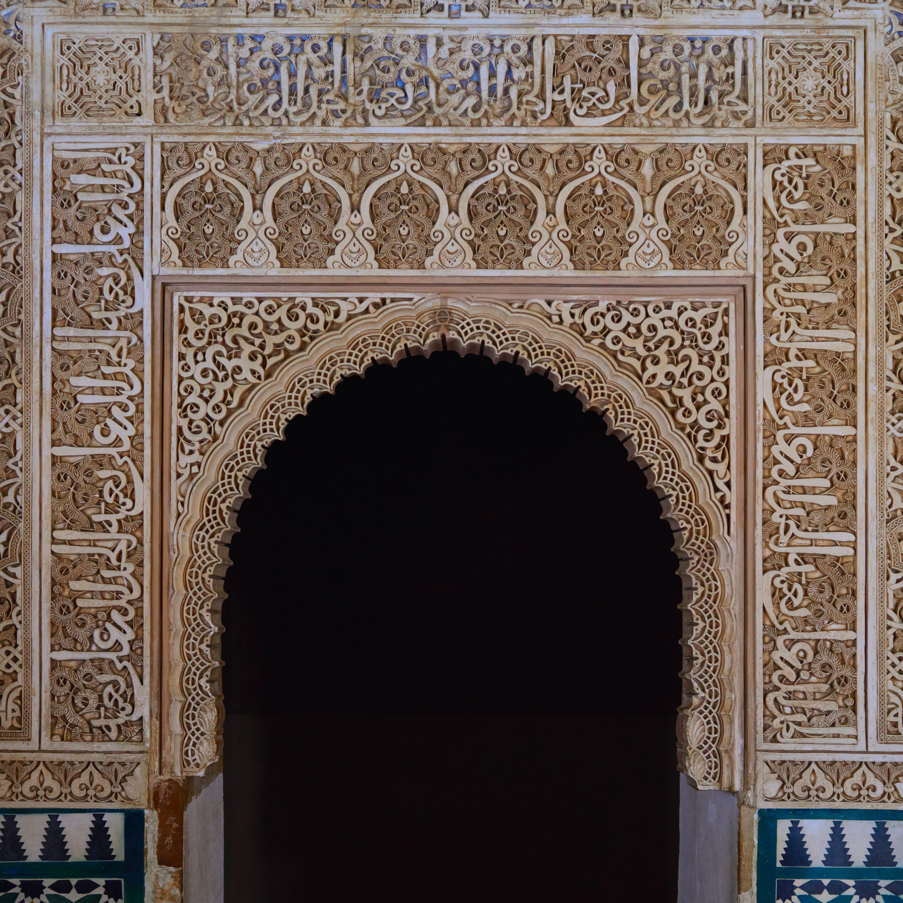 An intricate archway from inside the nasrid palace. The archway is pure darkness, but the surround is filled with intricate hand carved patterns in the stone. There are remnants of a deep blue paint that previously filled the engravings.
