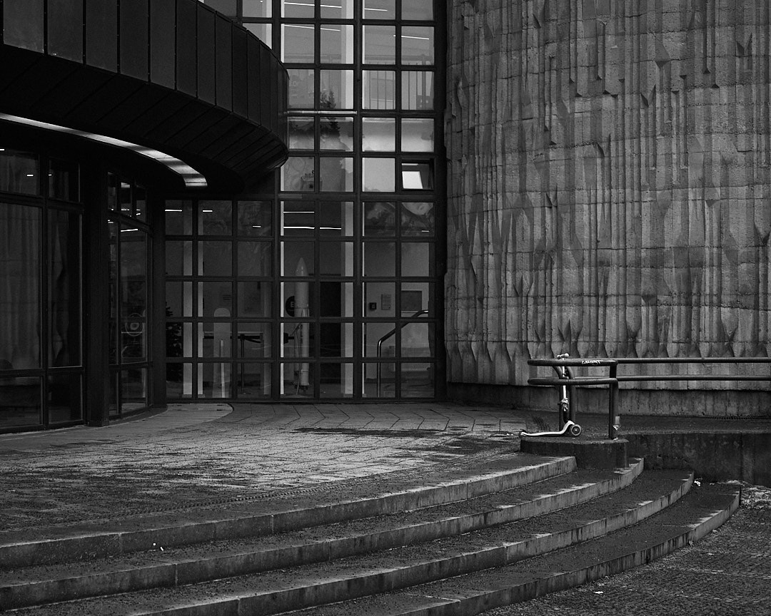 A black and white scene of the entrance to a building. In the foreground are curved steps, leading to a fence and an abandoned childrens scooter. In front of the entrance is a white gravelly texture, and to the side a lattice of windows with a view of empty halls.