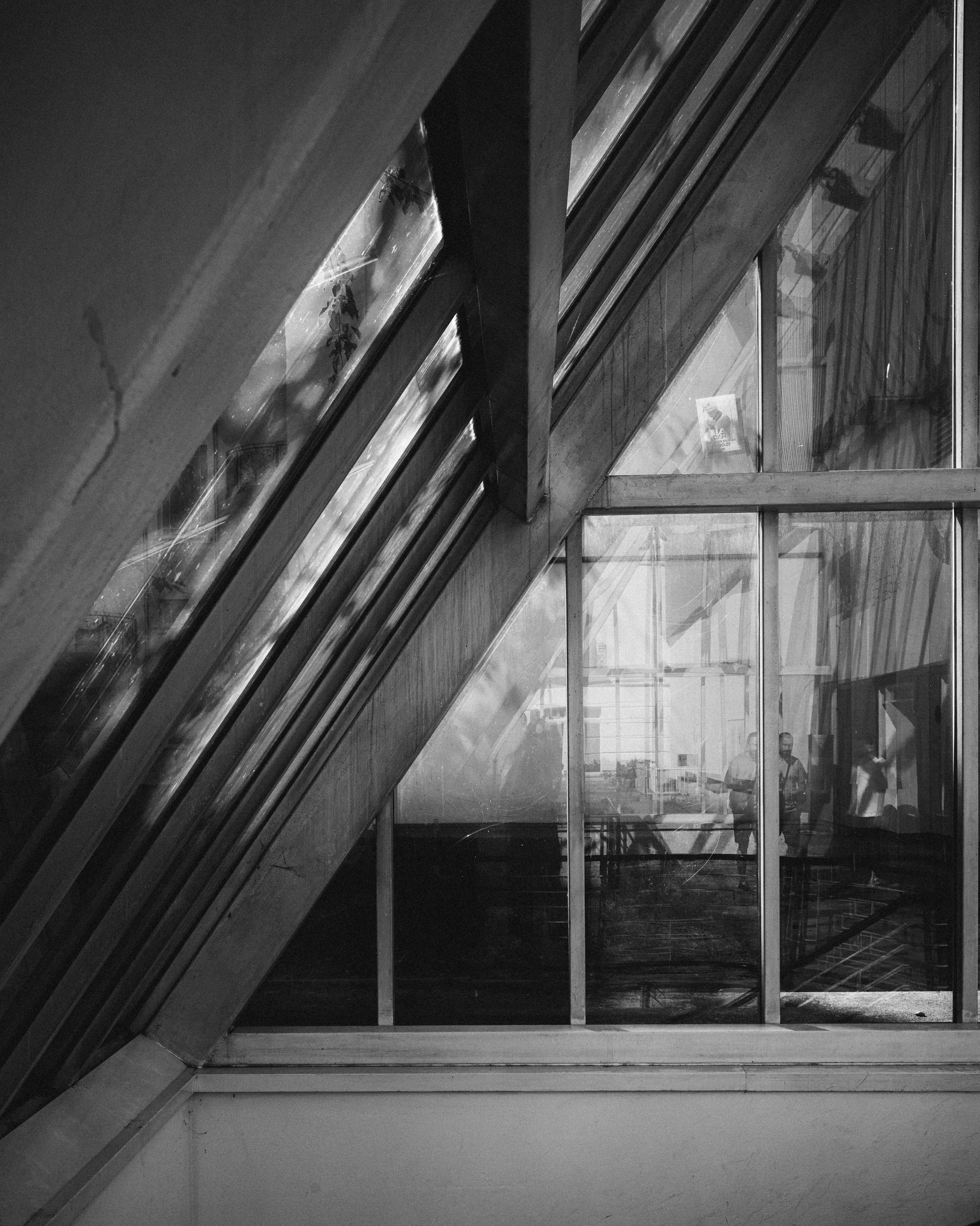 A black and white photograph of an angled window. The primary window runs at an angle from the top right to the bottom left of the frame. The windows are scratched and dirty. Their are many reflections of people walking, other buildings and benches. The window to the left that acts as a roof has patterns caused by the falling of light through trees. It invokes a slightly uncomfortable feeling through the grunginess.