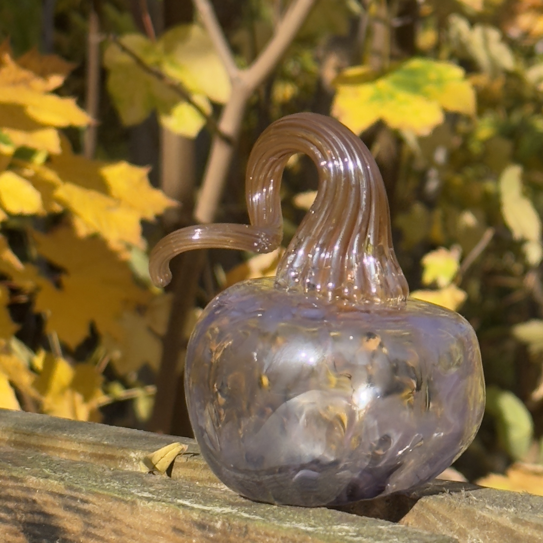 A purple glass pumpkin sat on a piece of wood. Yellow leaves are in the background.