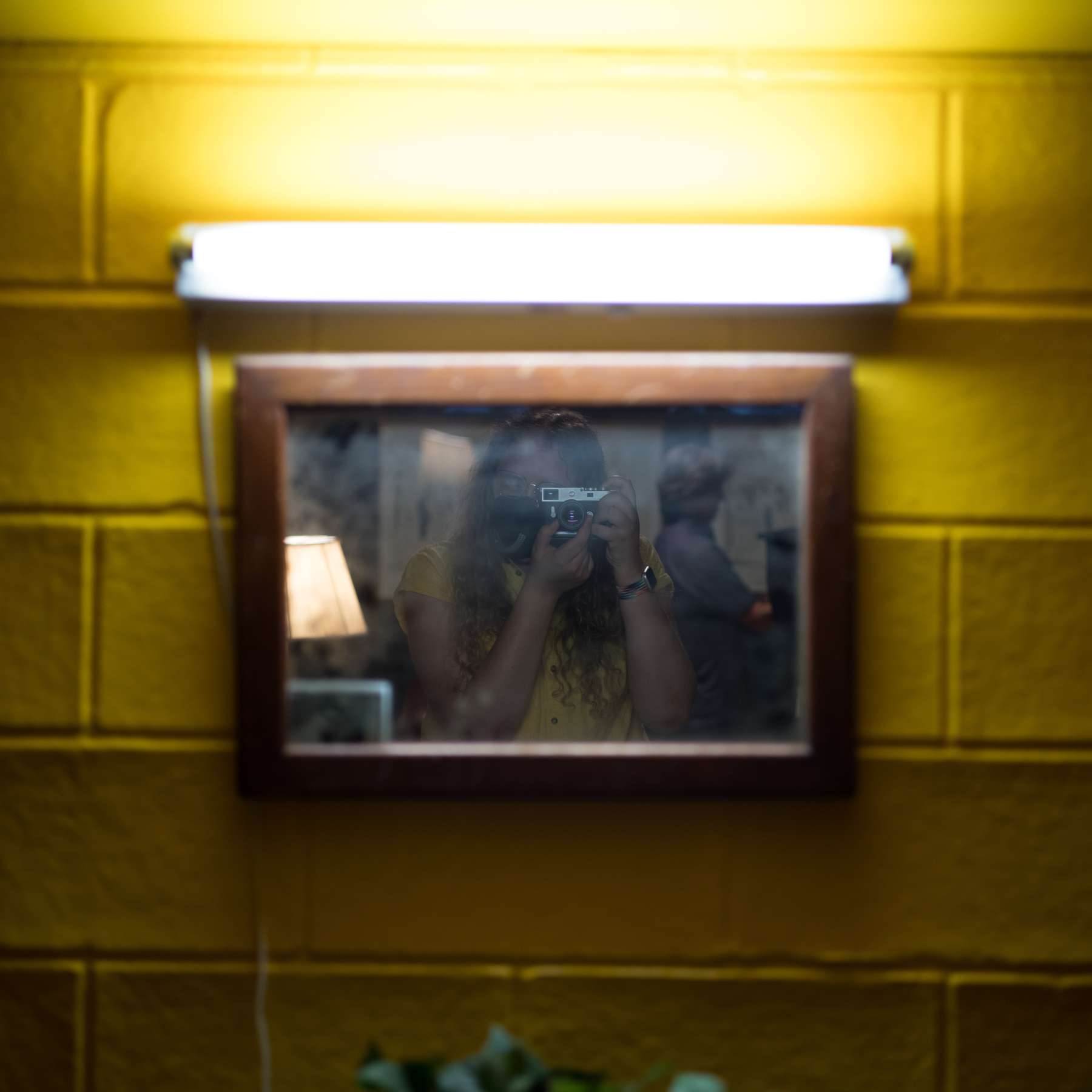 A yellow brick wall is adorned with a small dirty mirror and above it sits a bright tube light. In the mirror you can see a girl in a yellow shirt taking a photo directly into the mirror.