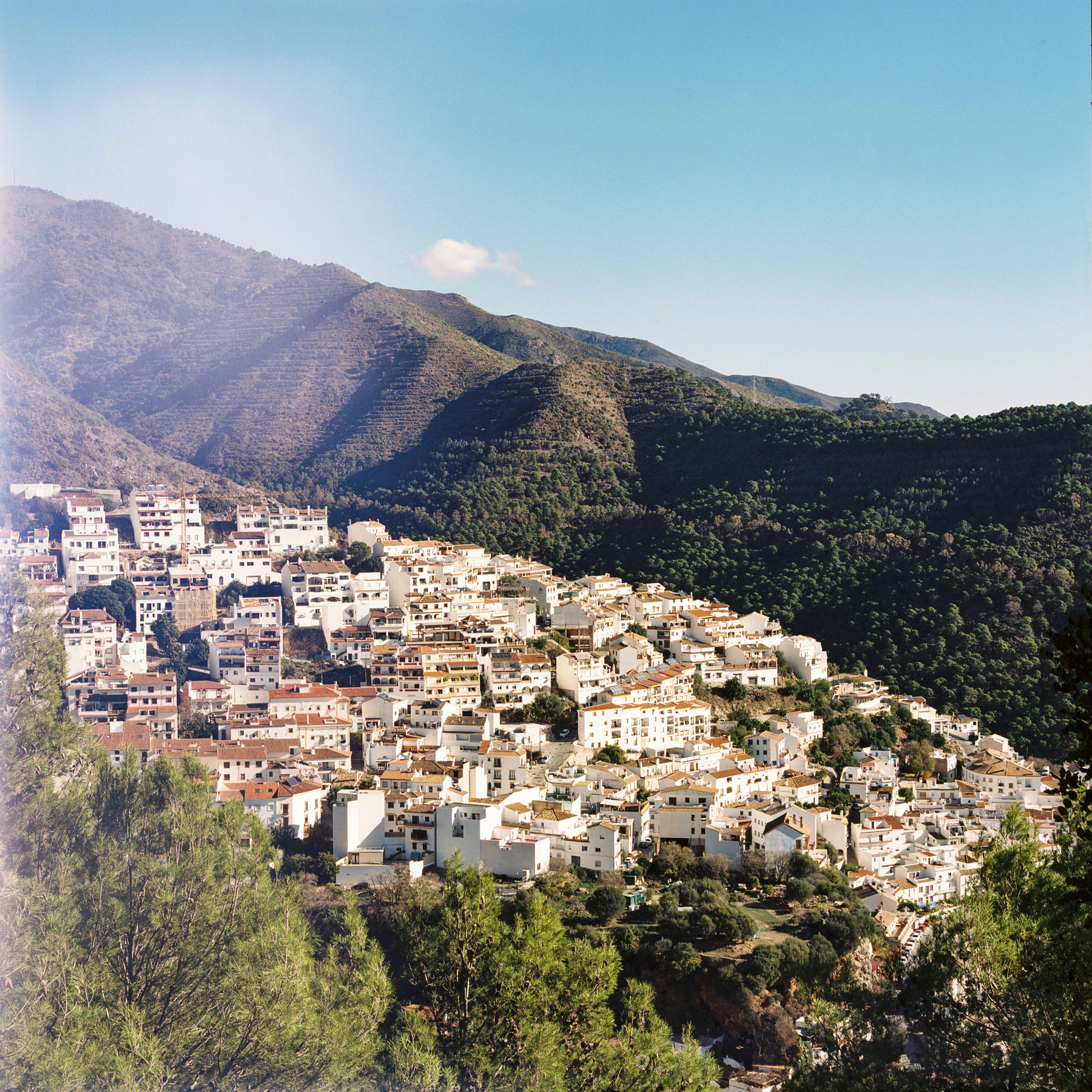 A photograph overlooking a town of white buildings and terracotta roofs. The town is backed by a tree covered mountain side. Above the mountain is a blue sky with hints of wispy cloud. To the left of the frame are some like leaks that distort color.
