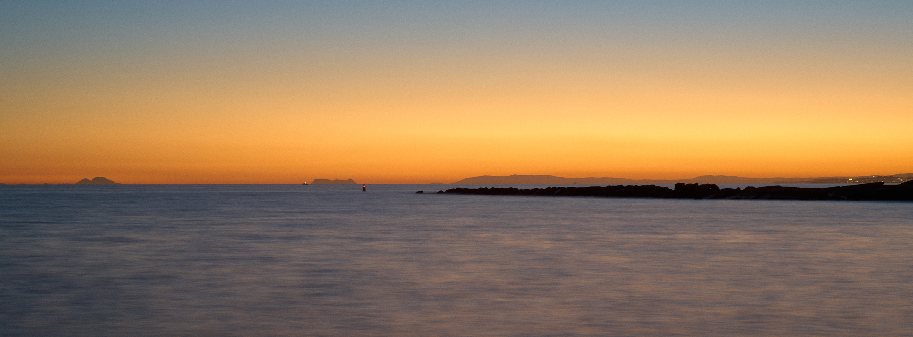 A sunset over Spain, Gibraltar, and Ceuta. The sea is rough but painterly, with swashes of blues and whites. The sky is a gradient of deep orange on the horizon going through yellow to blue hour hues.