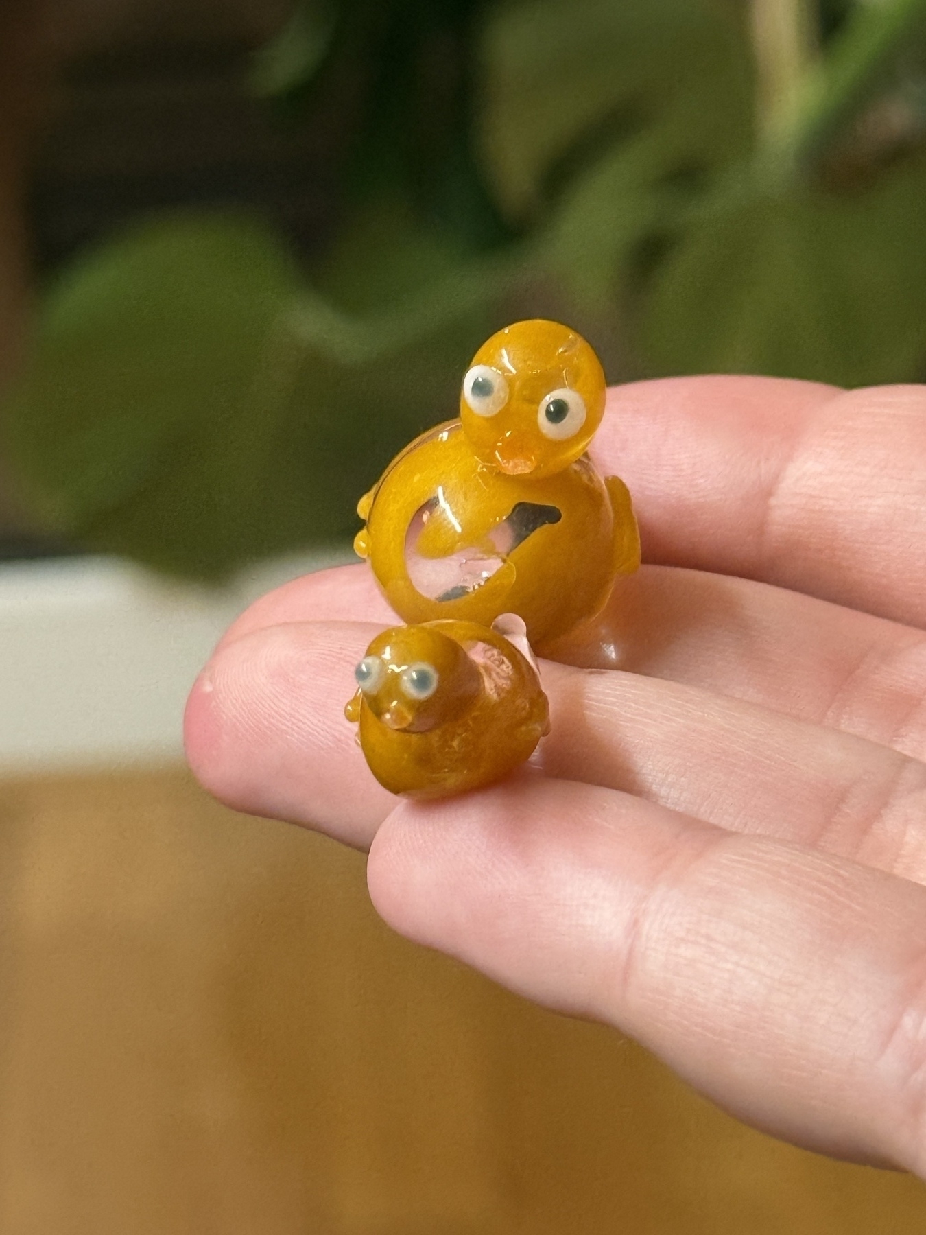 Two glass ducks, one smaller than the other, sat on someone’s fingertips, there is a plant in the background.